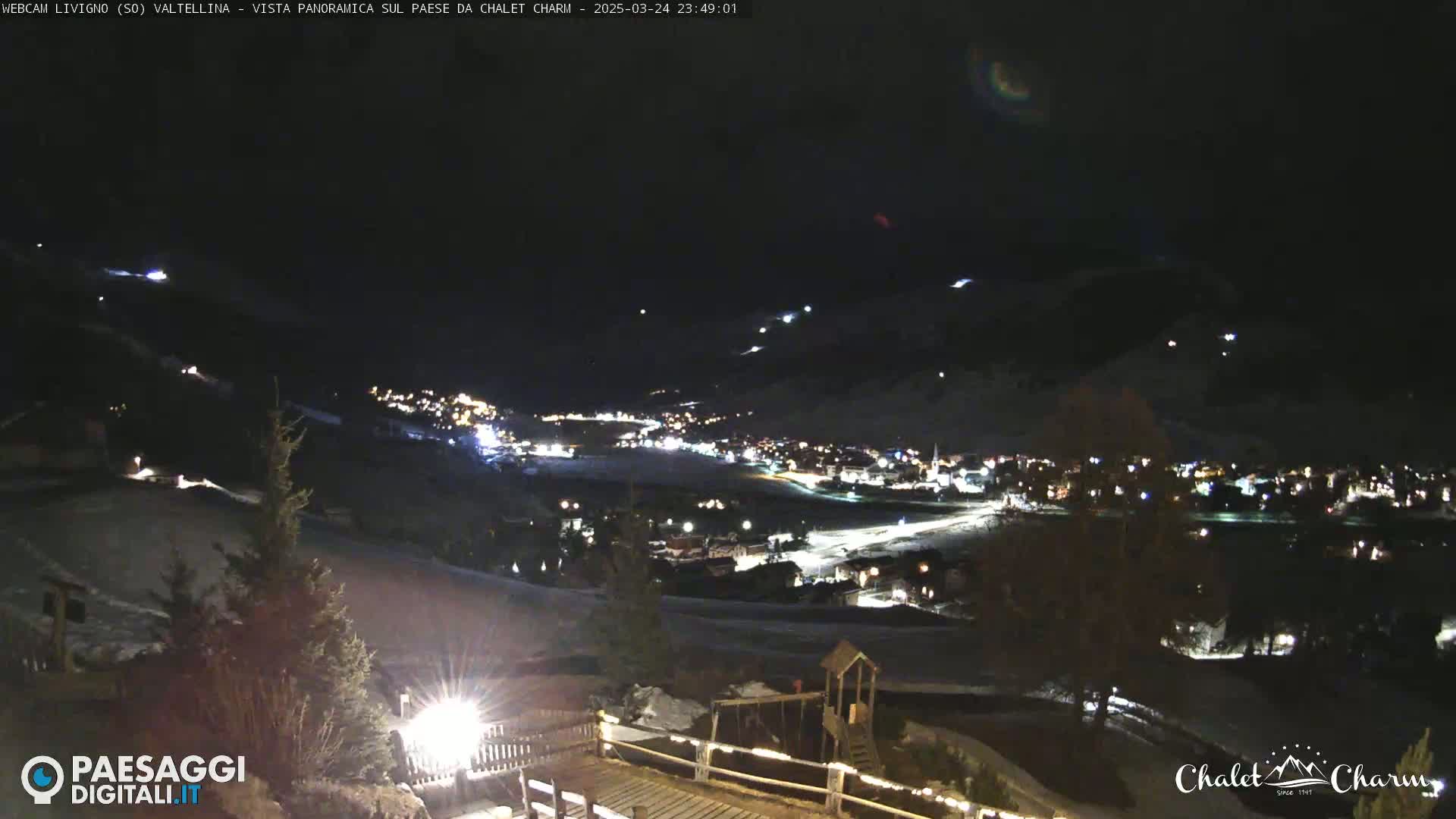 A nighttime panoramic view of a snow-covered village illuminated by lights, with a playground visible in the foreground under a clear, dark sky.