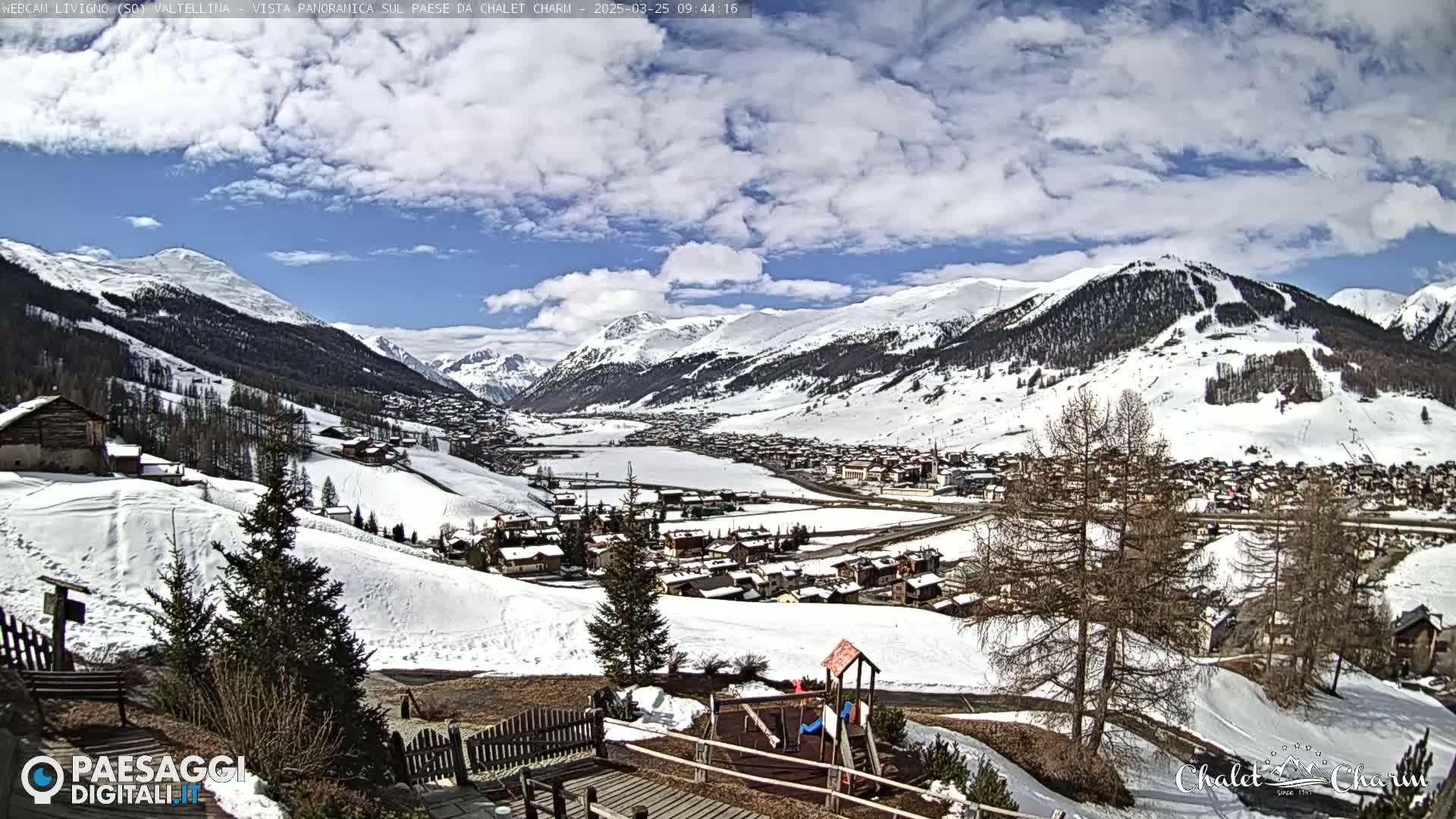 A snow-covered mountain village is viewed from a hillside on a partly cloudy day, with a playground visible in the foreground.