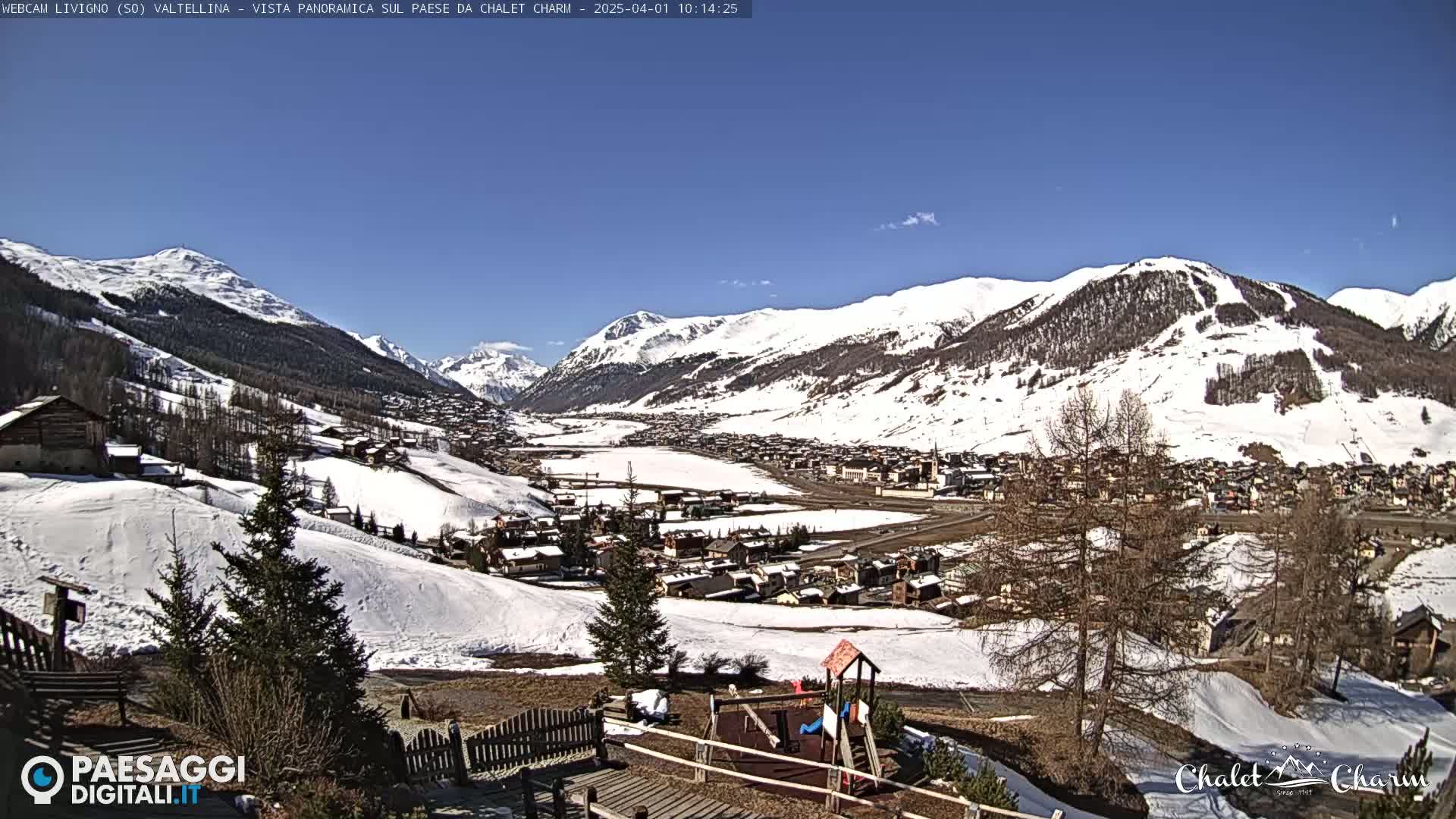 A sunny, snow-covered mountain valley town is viewed from a hillside, with a children's playground in the foreground.