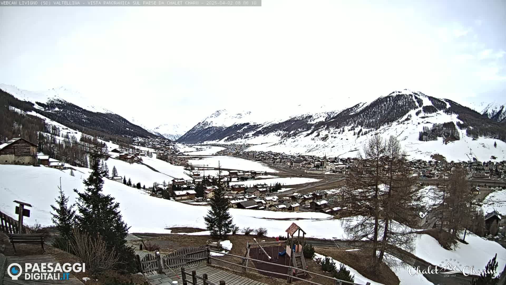 A snow-covered mountain valley town is viewed from a hilltop, with a playground visible in the foreground under an overcast sky.