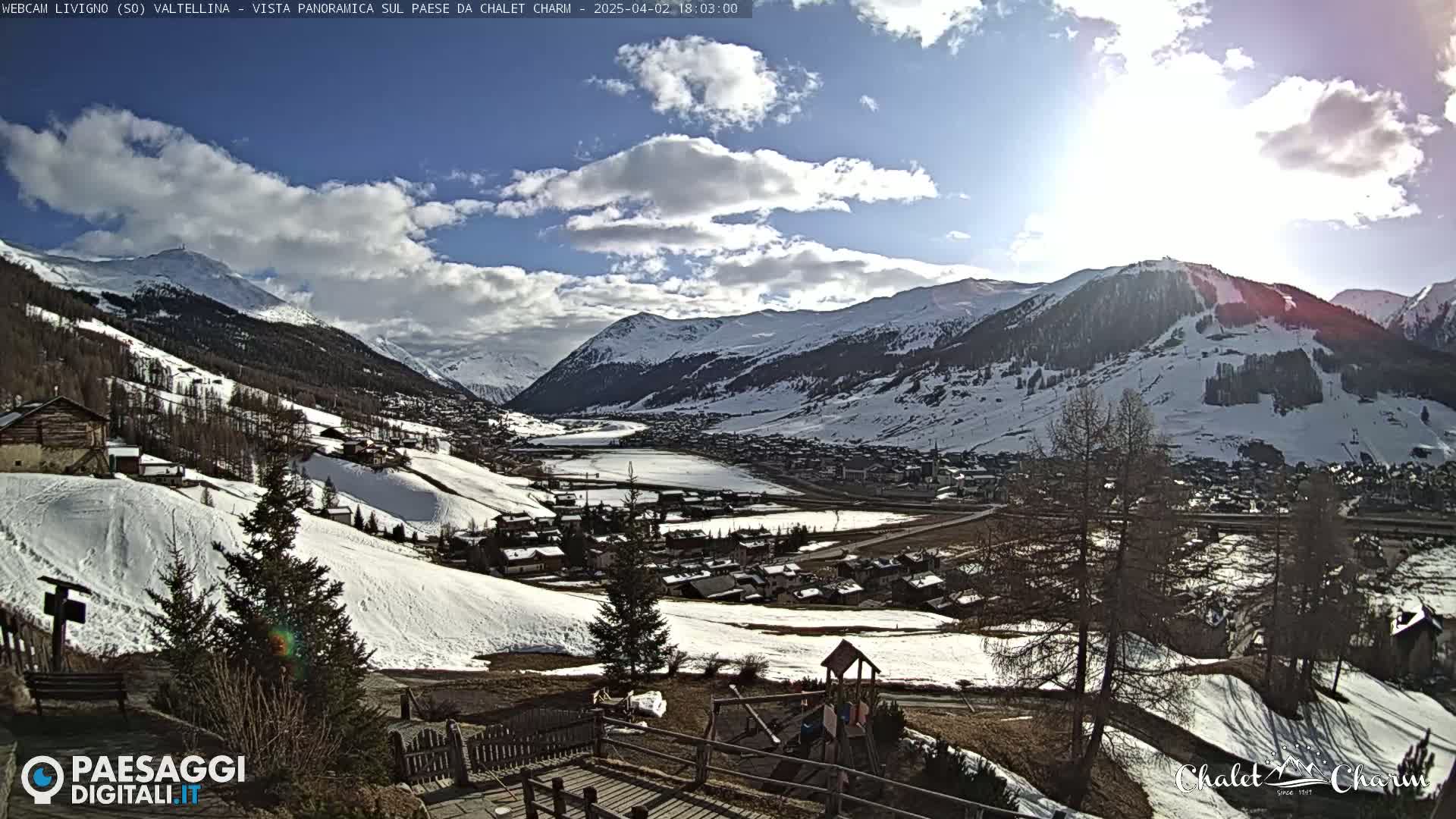 A sunny day with partly cloudy skies overlooks a snow-covered mountain valley town with a playground visible in the foreground.