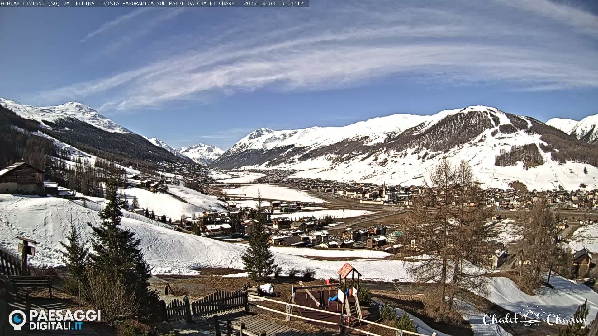 A snow-covered mountain valley town is viewed from a hilltop on a sunny day with a few wispy clouds.