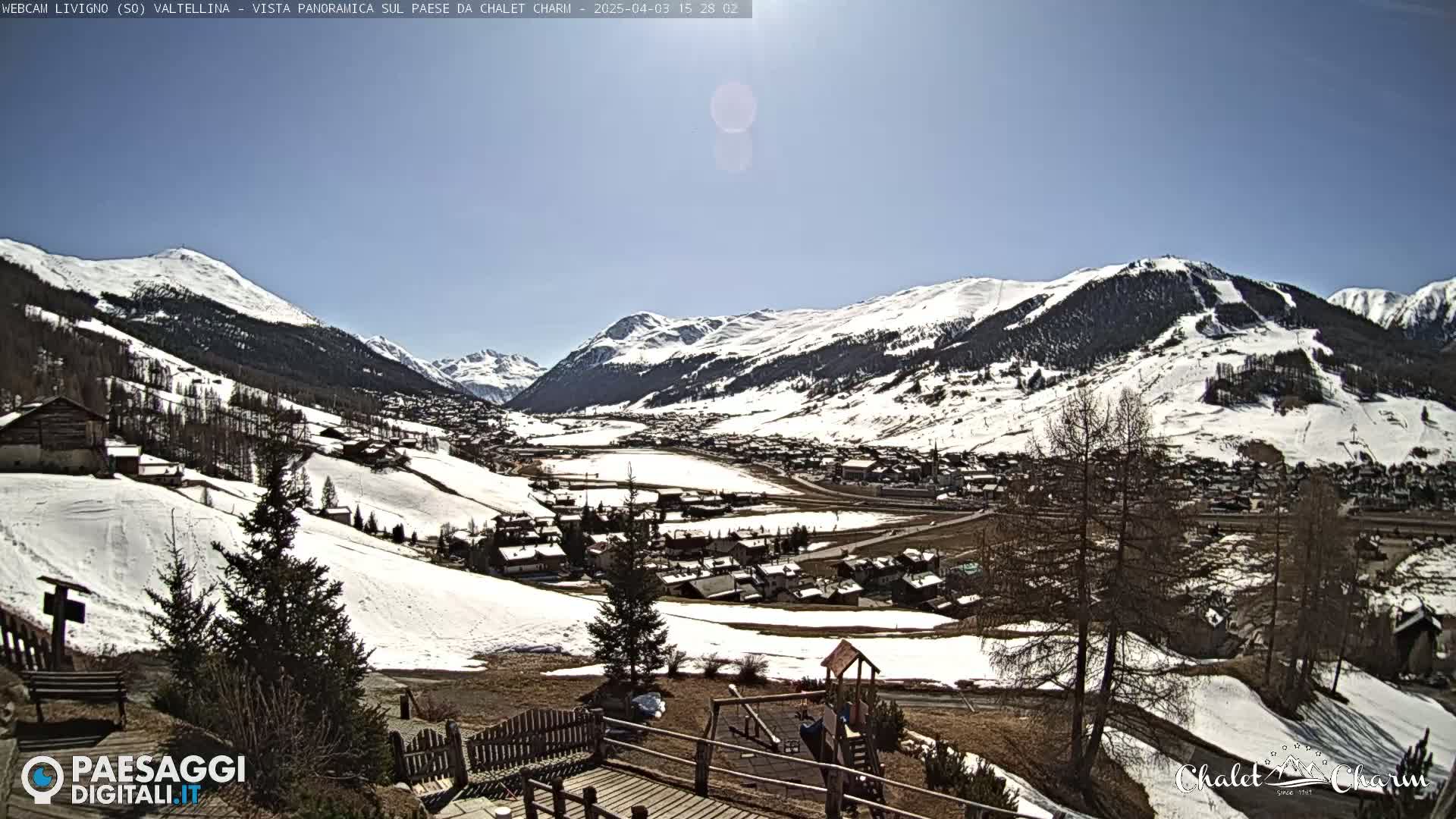 A sunny day reveals a snow-dusted mountain valley village, with patches of snow remaining on the hillsides and surrounding mountains.