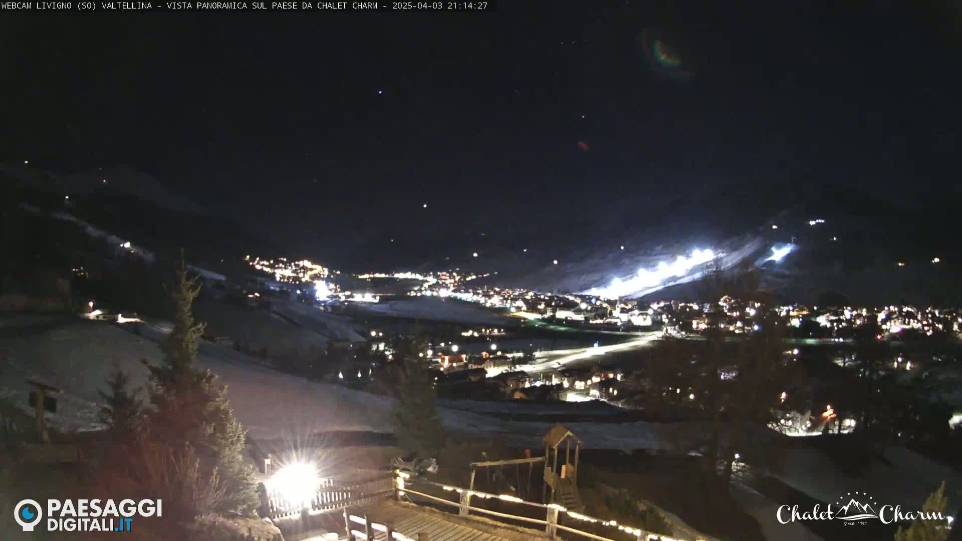 A nighttime view of a snow-covered town in a valley, illuminated by streetlights and lights on a nearby ski slope, under a clear, starry sky.