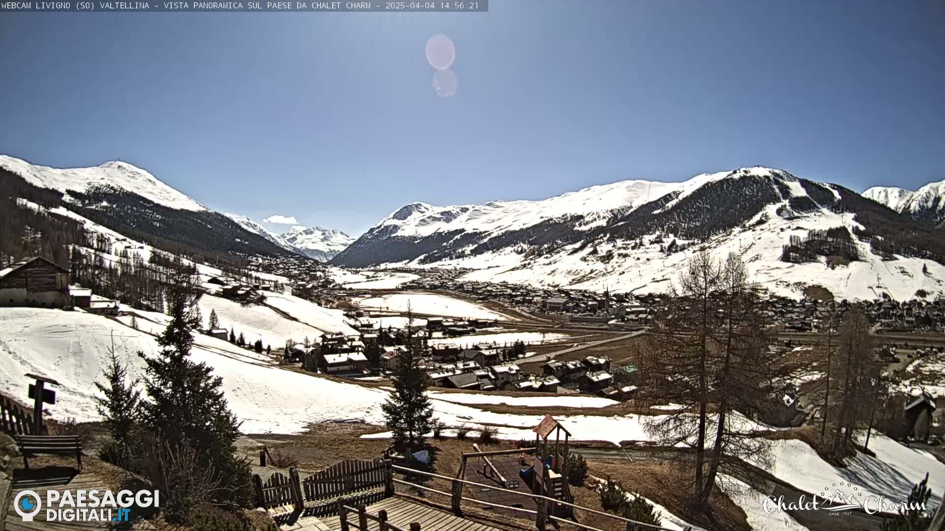 A sunny, snow-dusted mountain valley town is viewed from an elevated vantage point, with a playground visible in the foreground.
