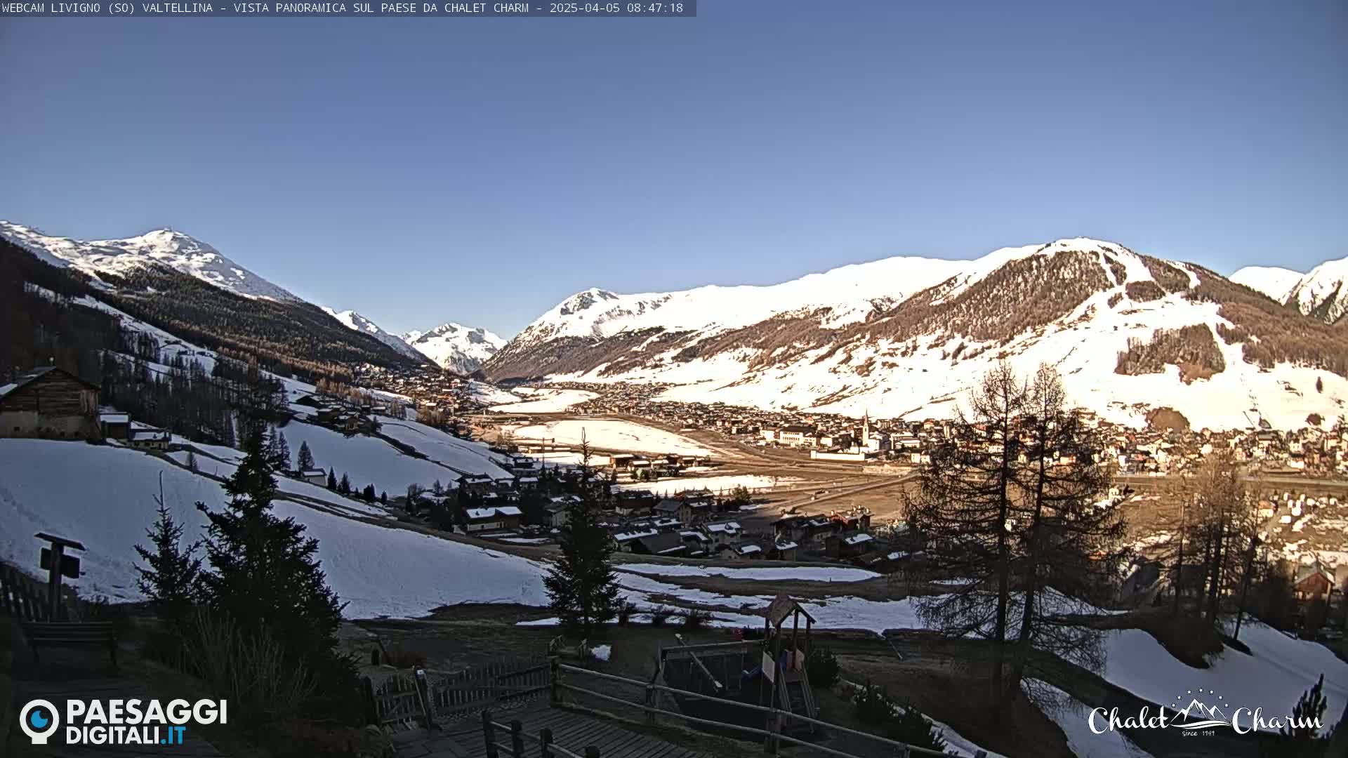 A panoramic view of a snow-dusted town nestled in a valley surrounded by snow-capped mountains under a clear blue sky.