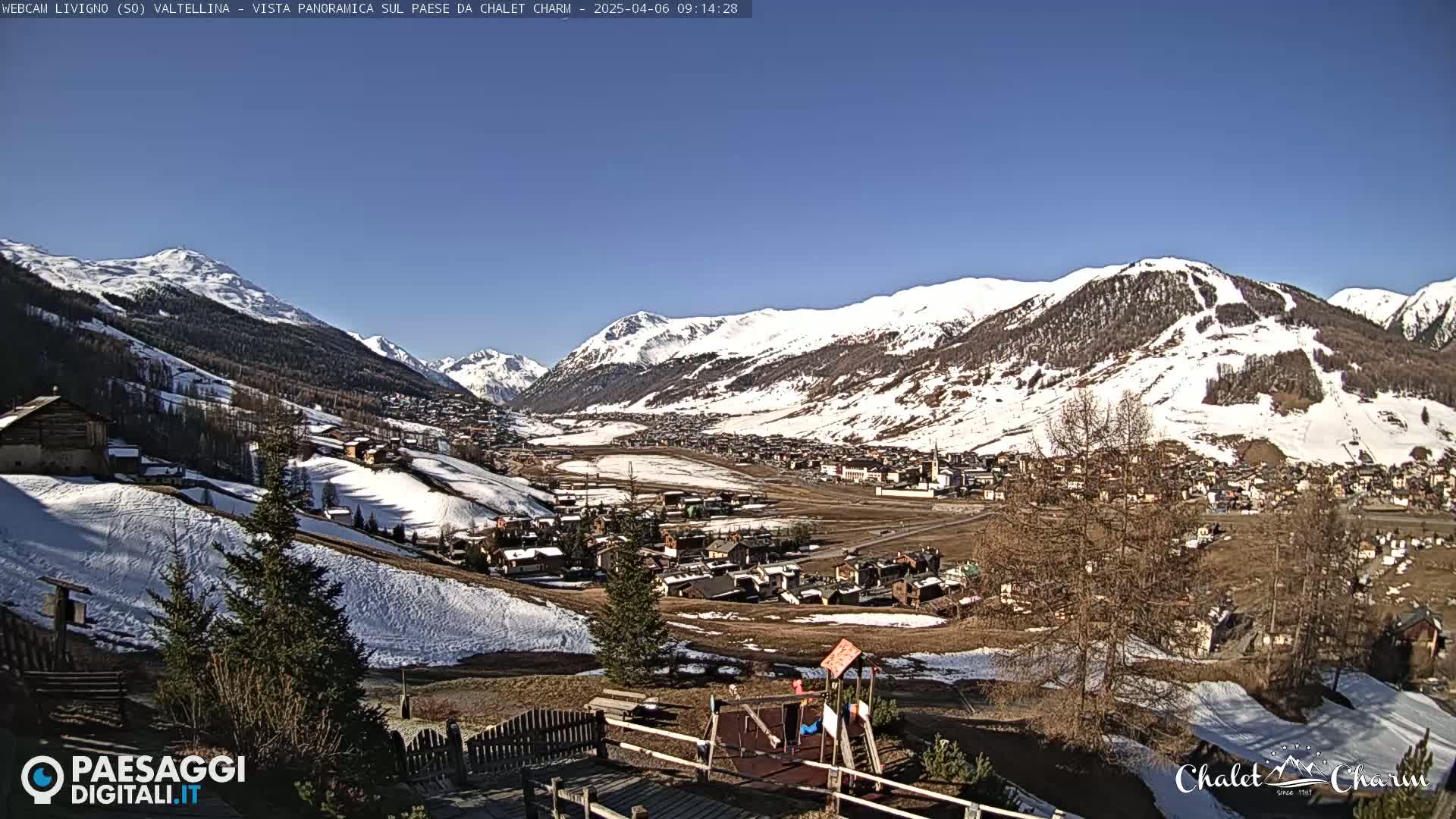 A sunny, snow-covered mountain valley town is viewed from a hillside, with a small playground visible in the foreground.