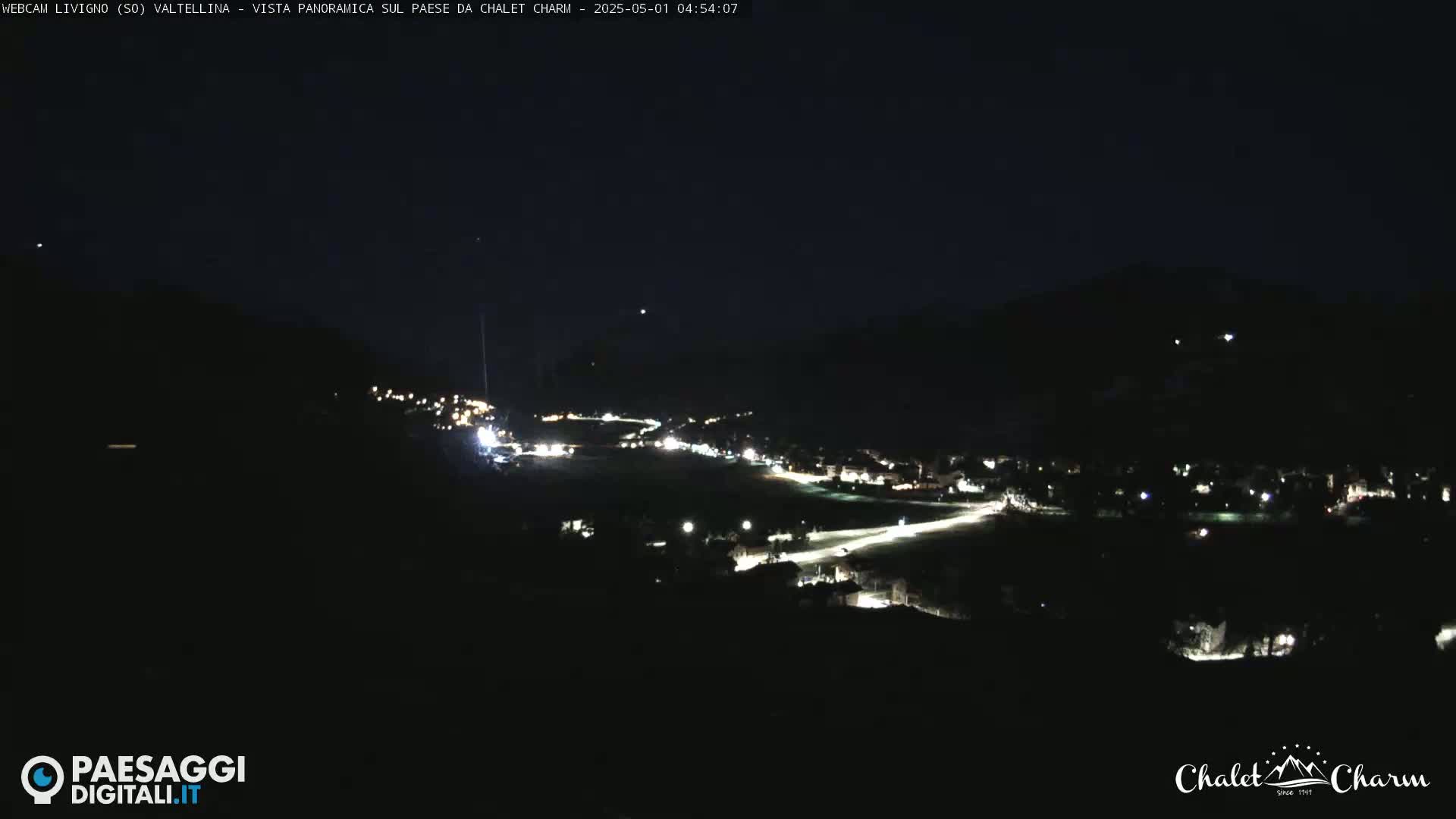 A nighttime, panoramic view of a town nestled in a valley, illuminated by scattered lights.