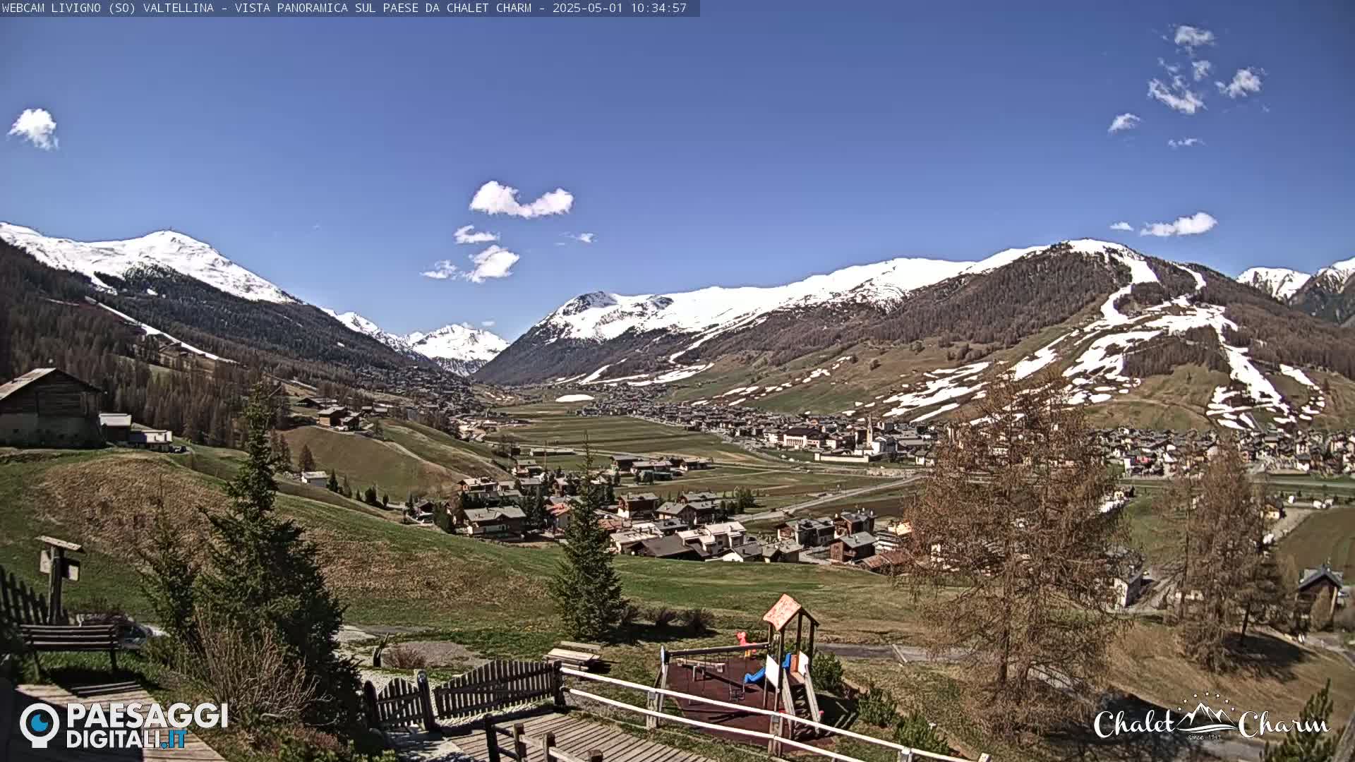 A sunny day reveals a picturesque mountain valley town nestled amongst snow-capped peaks, with a playground visible in the foreground.