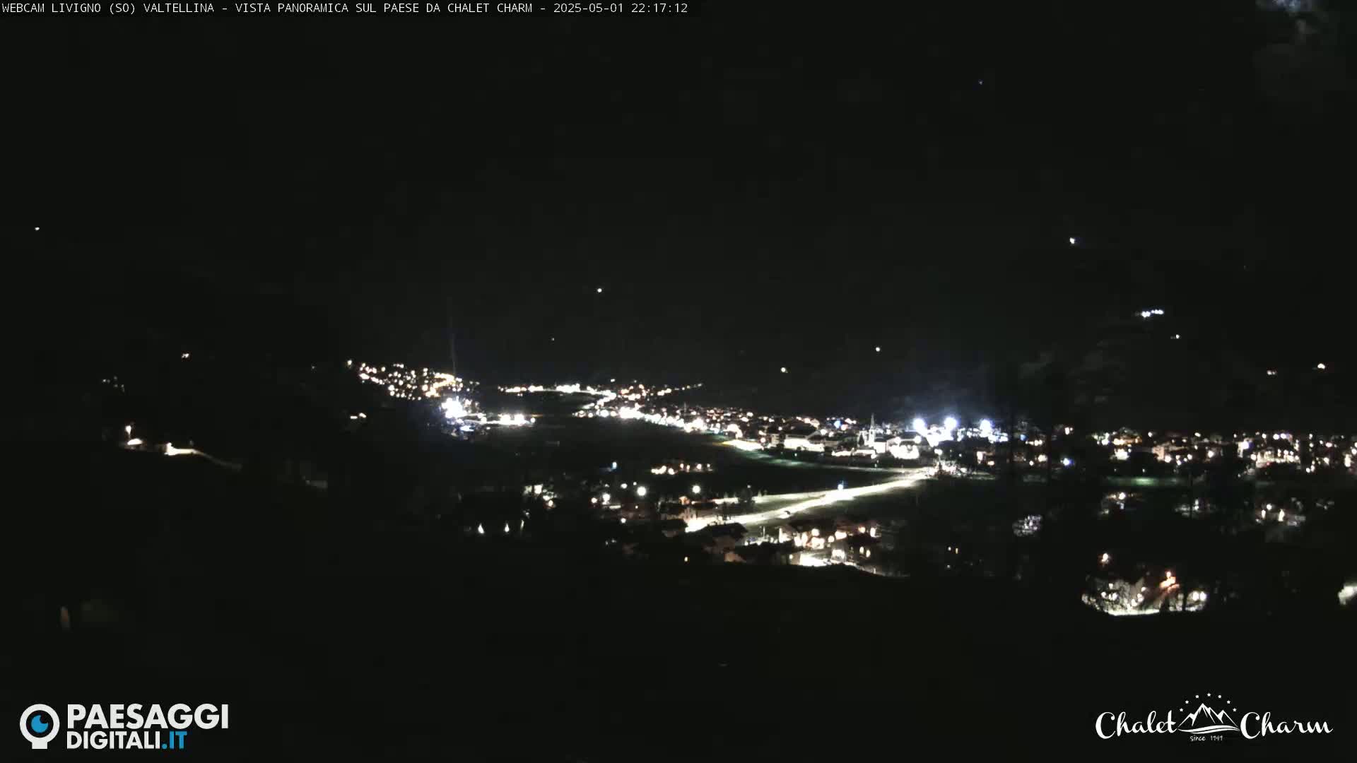 A nighttime, panoramic view of a town nestled in a valley, illuminated by scattered lights under a dark, clear sky.