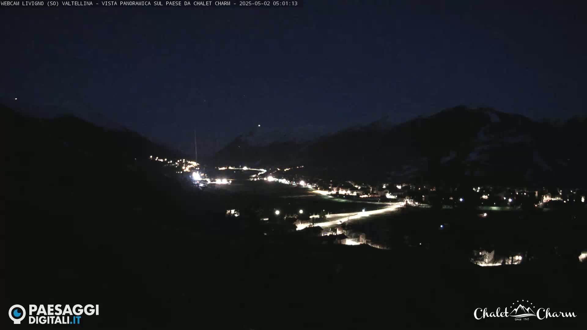 At night, a village nestled in a valley between dark mountains is illuminated by scattered lights.