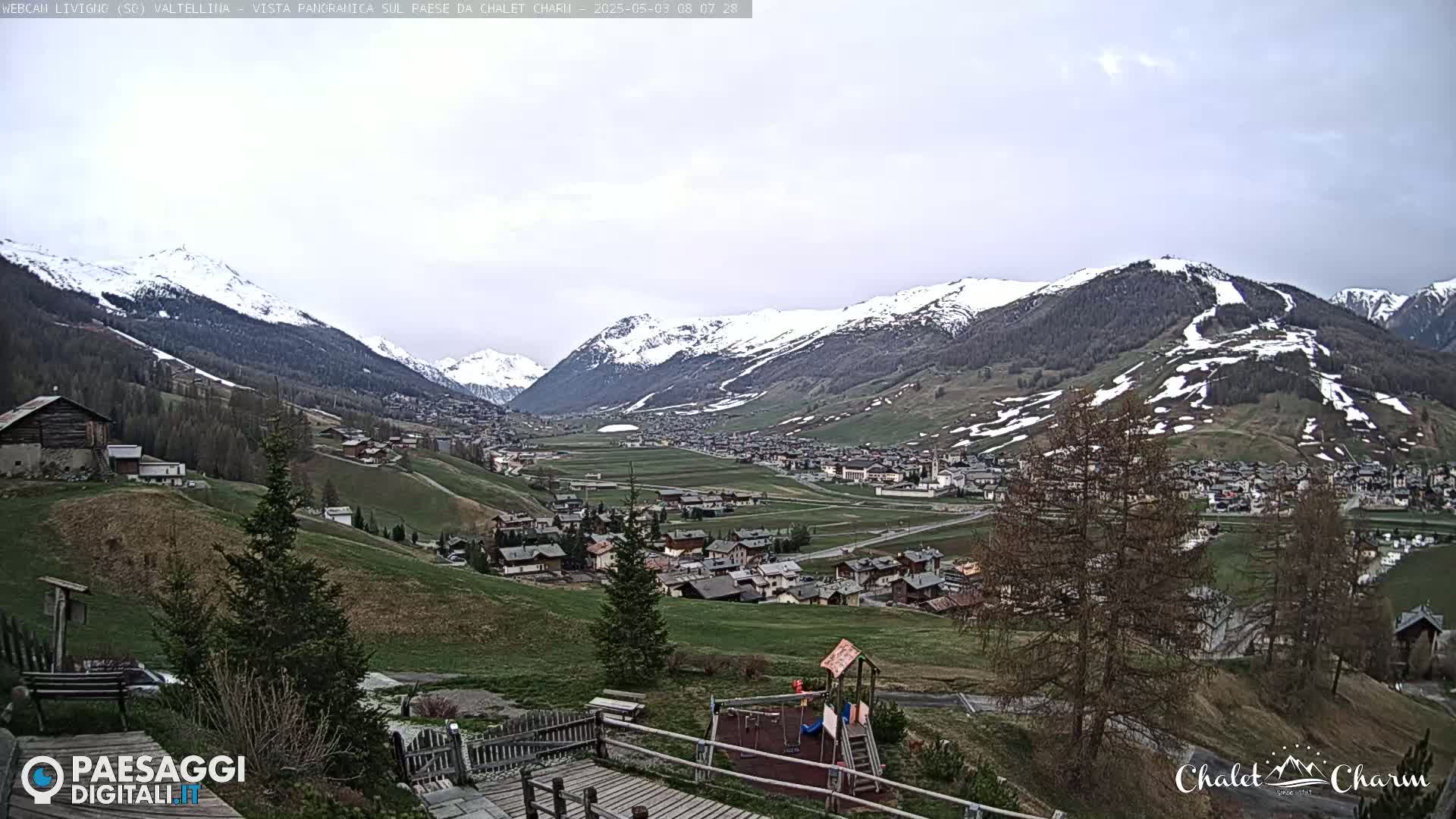 A view of a village nestled in a valley between snow-capped mountains under an overcast sky.