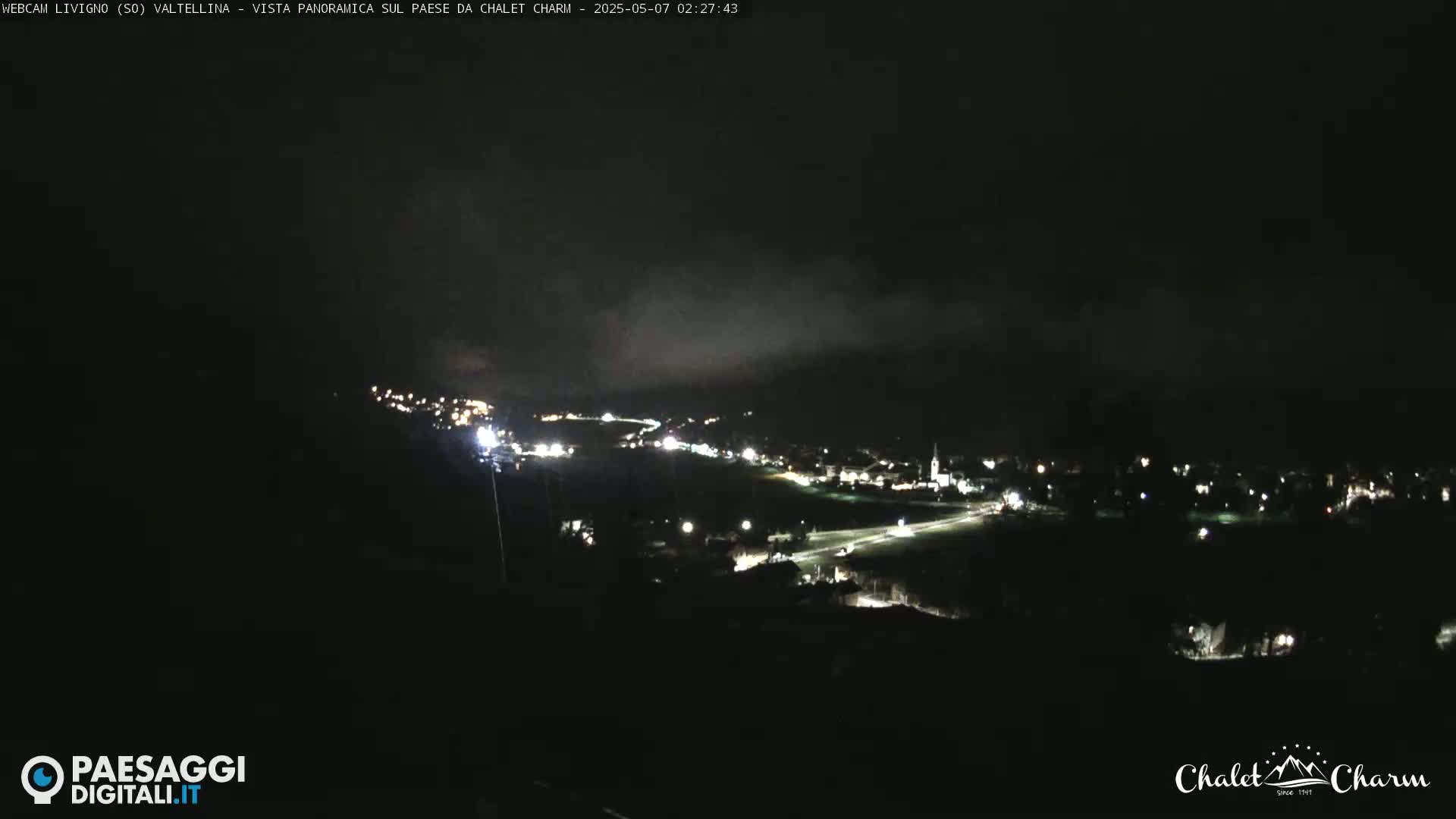 A nighttime long shot of a dimly lit town nestled in a valley under a dark, cloudy sky.