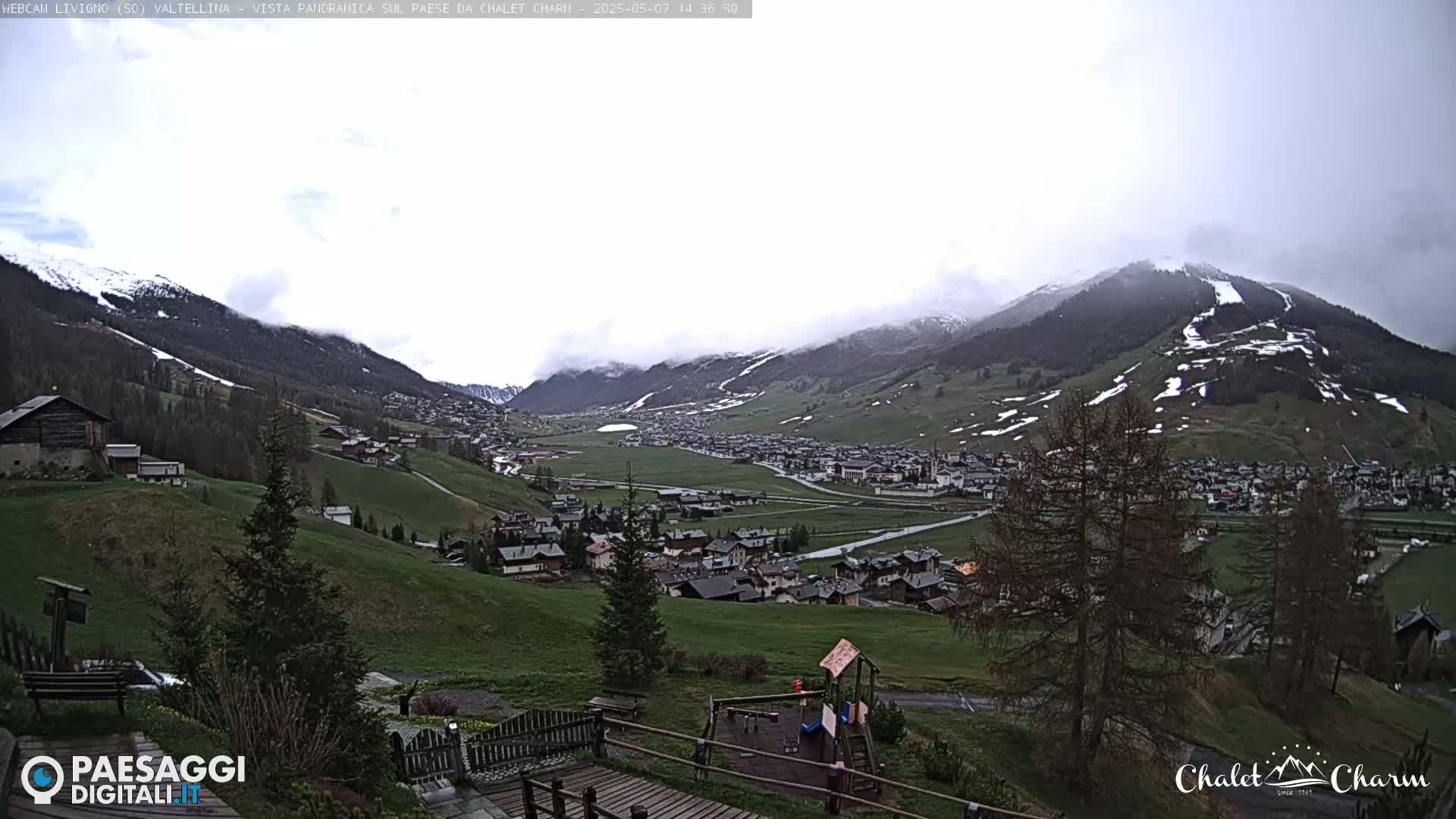 A mountain valley town is visible under an overcast sky, with patches of snow remaining on the surrounding mountains.