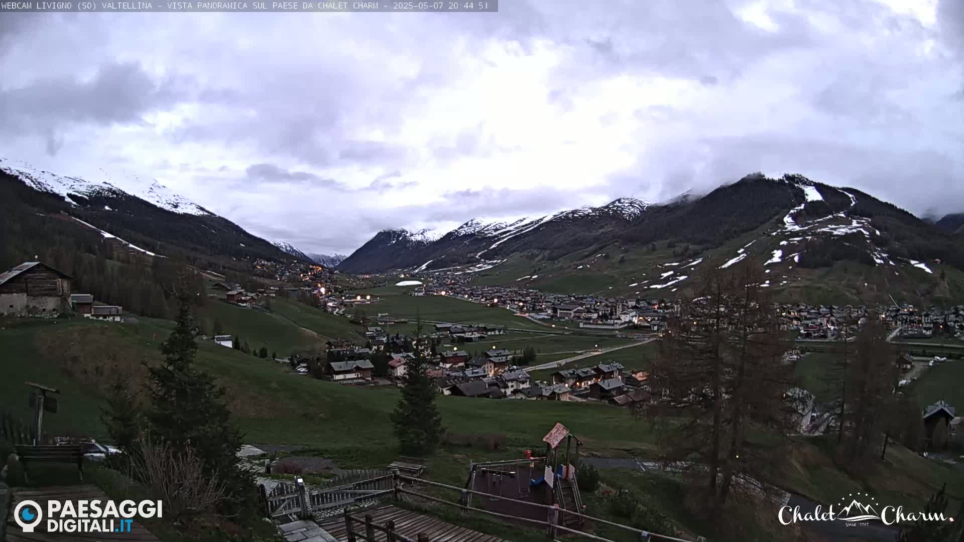 A village nestled in a valley between snow-capped mountains under a cloudy, twilight sky.