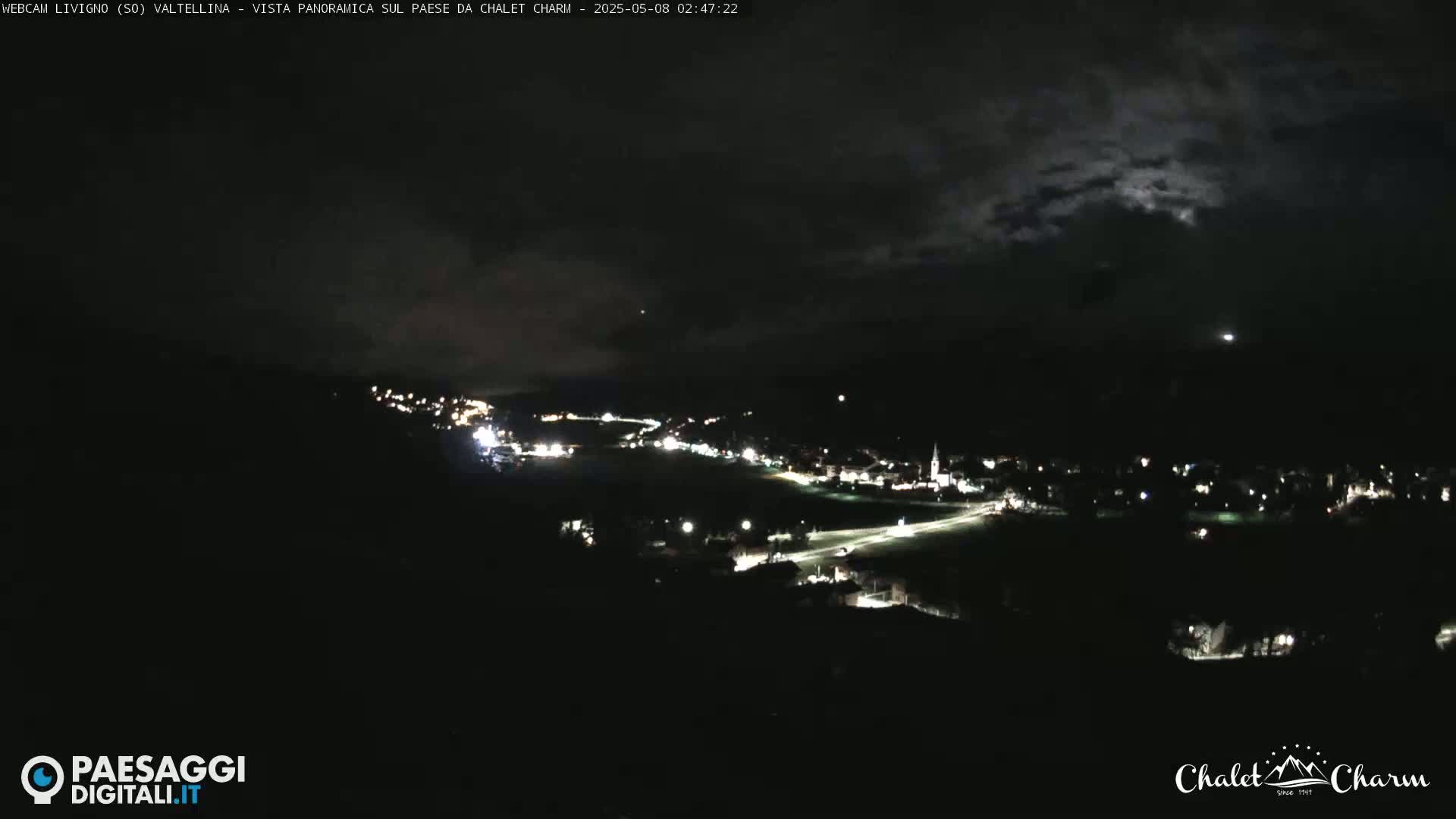 A nighttime panoramic view of a town illuminated by lights under a partly cloudy sky.