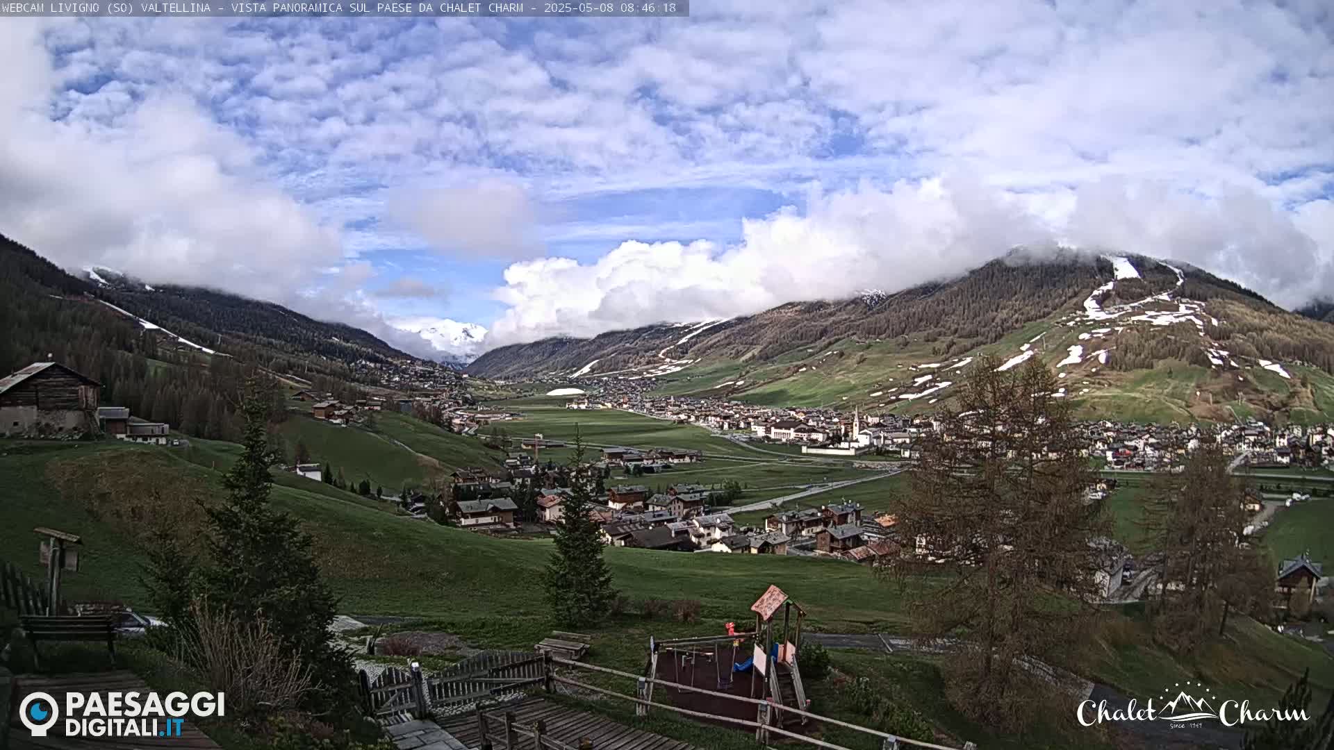 A partly cloudy day reveals a panoramic view of a valley town nestled among rolling green hills and snow-capped mountains.