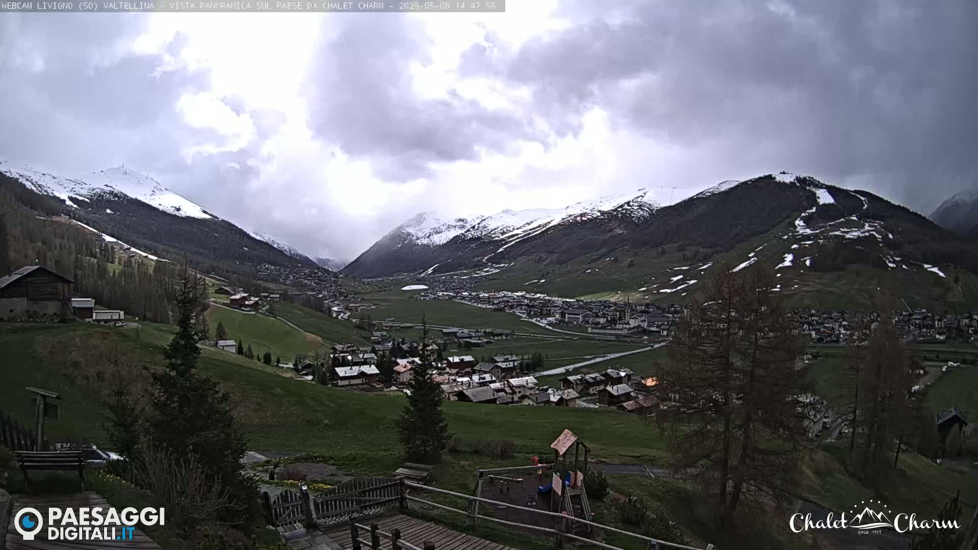 A panoramic view of a snow-capped mountain valley town under a cloudy sky shows green hillsides, a playground, and sparsely populated areas.