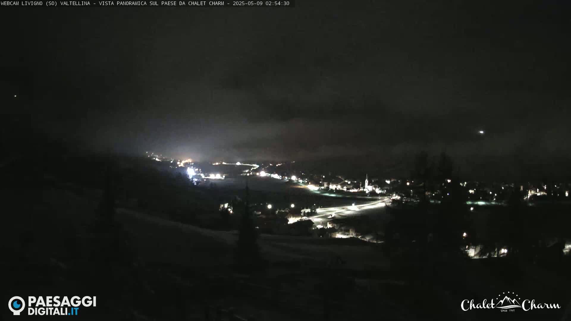 A nighttime, panoramic view of a snow-covered town illuminated by scattered lights under a dark, cloudy sky.
