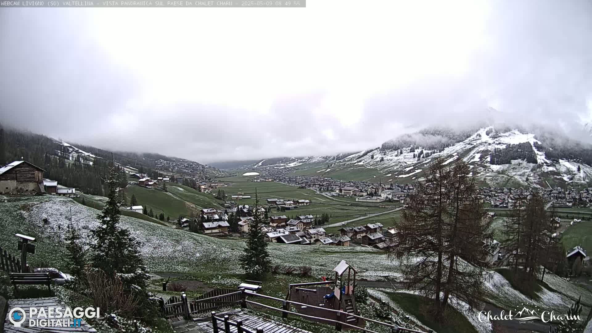 A snow-dusted mountain village nestled in a valley, under a cloudy sky with light snowfall.
