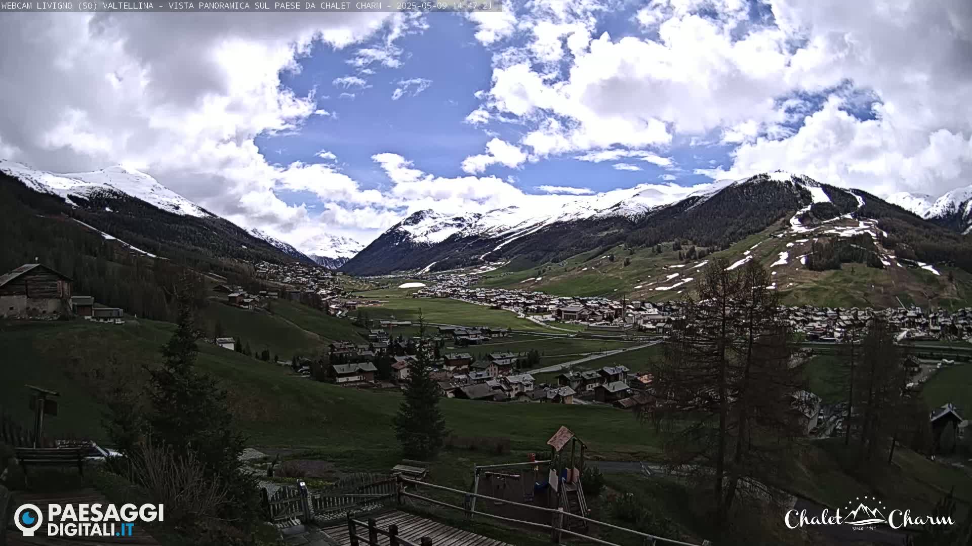 A partly cloudy day reveals a panoramic view of a snow-capped mountain valley village with a playground in the foreground.
