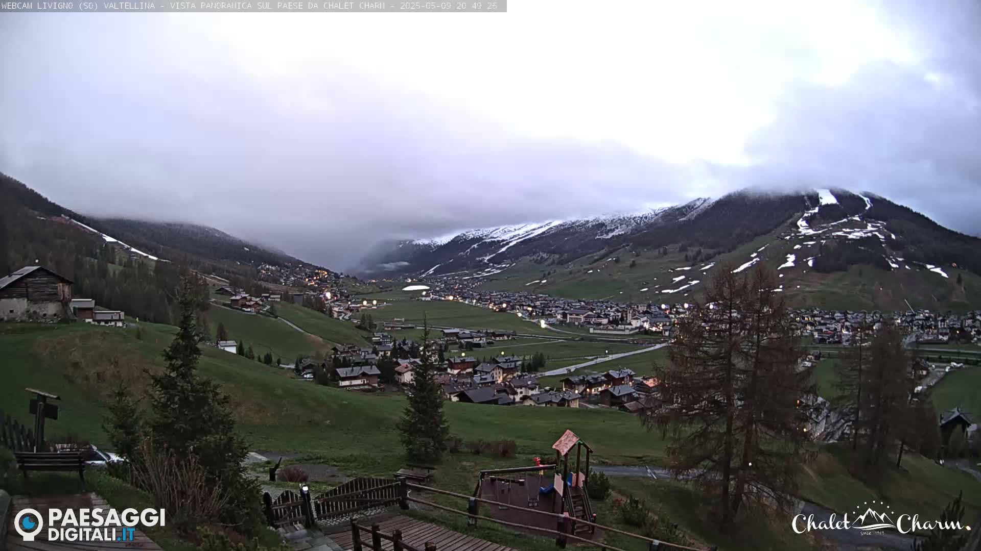 A mountain village nestled in a valley, partially shrouded in clouds, with snow-capped mountains in the background.