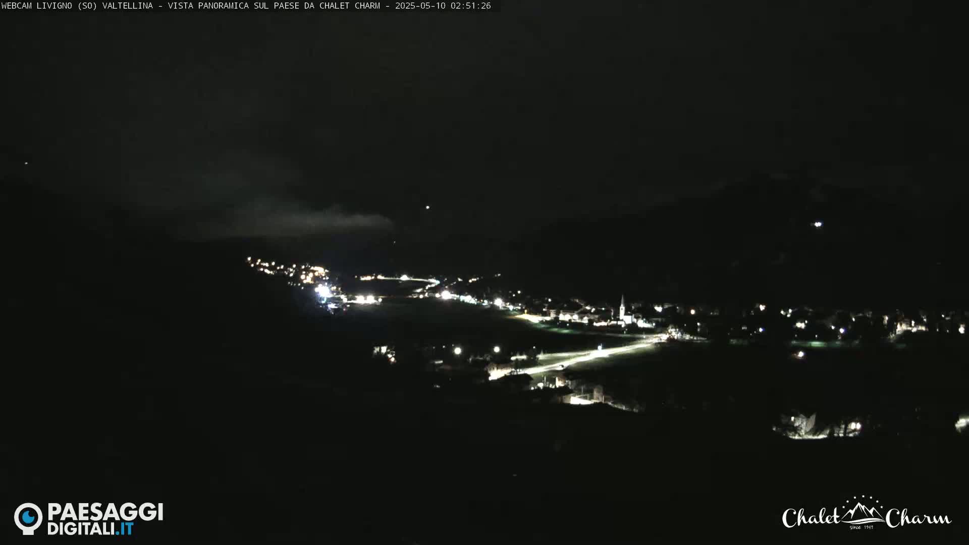 A nighttime, long-range view of a small, dimly lit town nestled in a valley, partially obscured by fog or low clouds.
