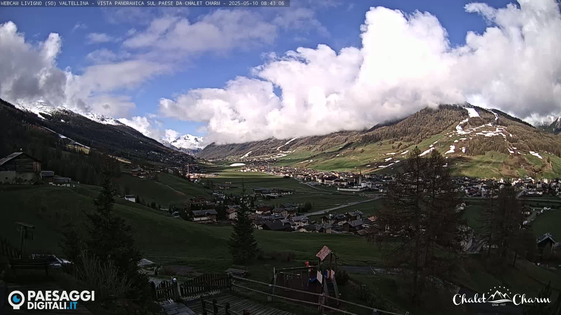 A partly cloudy day reveals a valley town nestled among snow-capped mountains and rolling green hills, with a playground visible in the foreground.