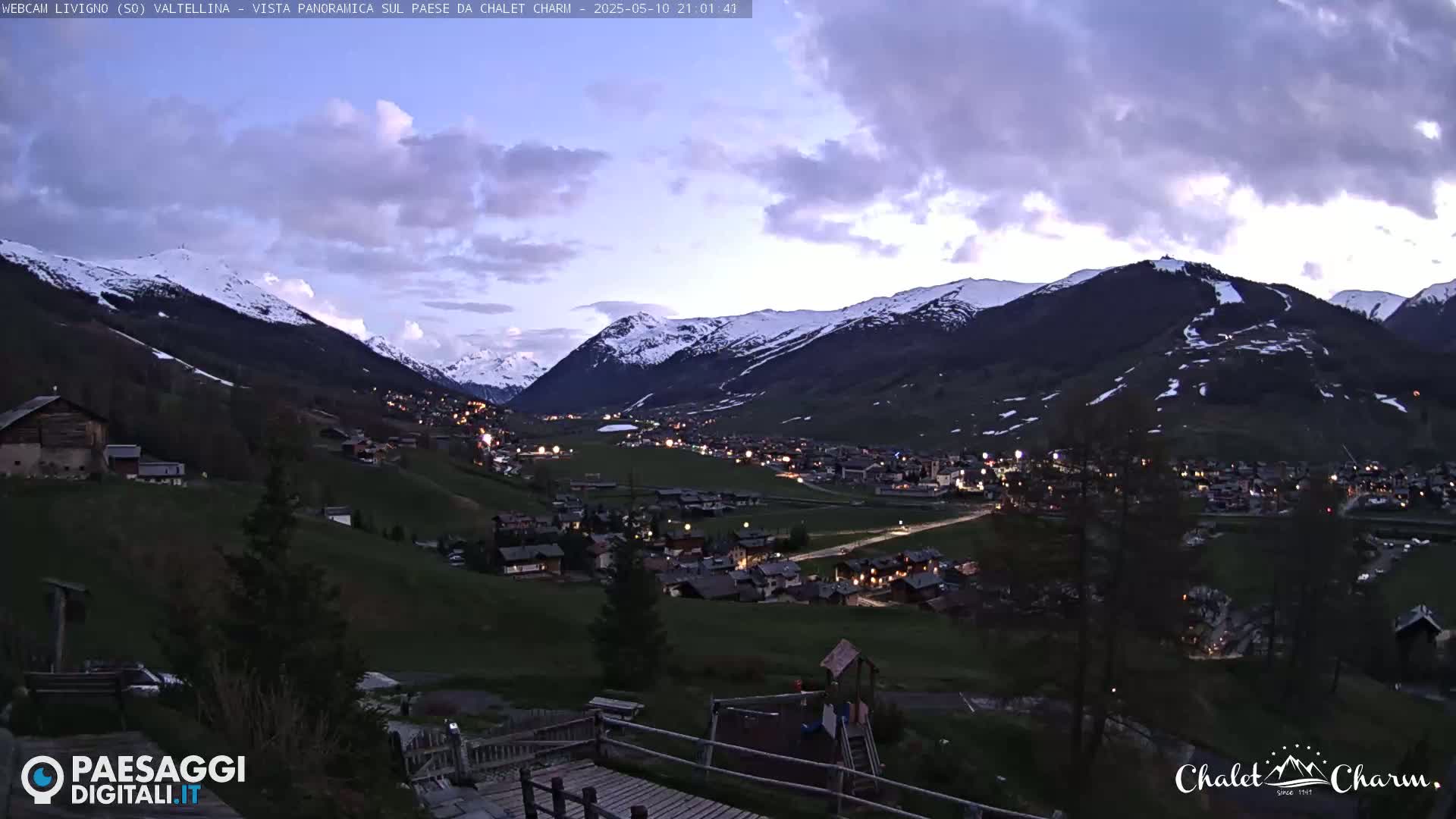 A twilight view of a snow-capped mountain valley with a village nestled below, under a partly cloudy sky.