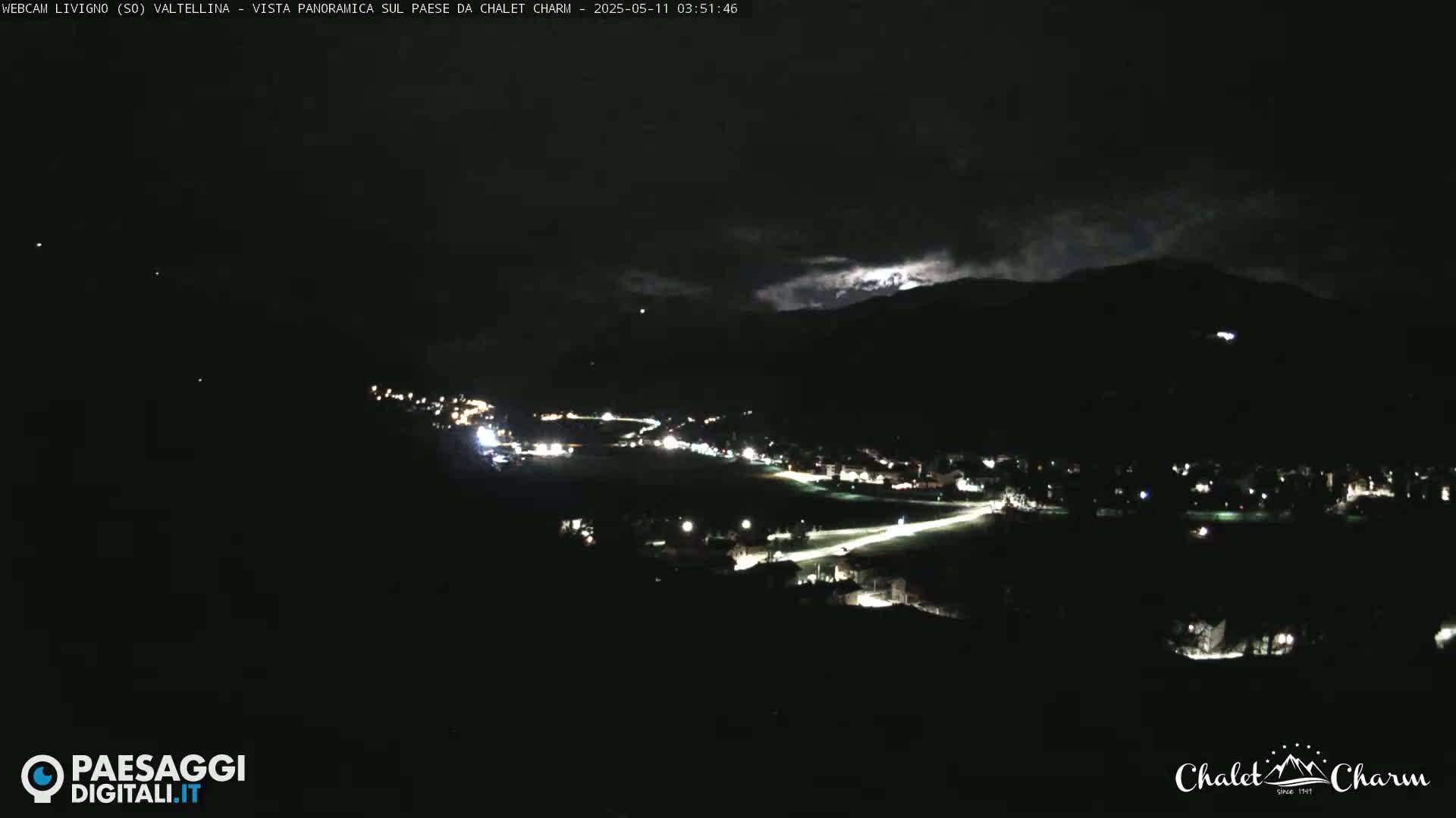 A nighttime, panoramic view of a town nestled in a valley beneath dark, cloudy mountains.