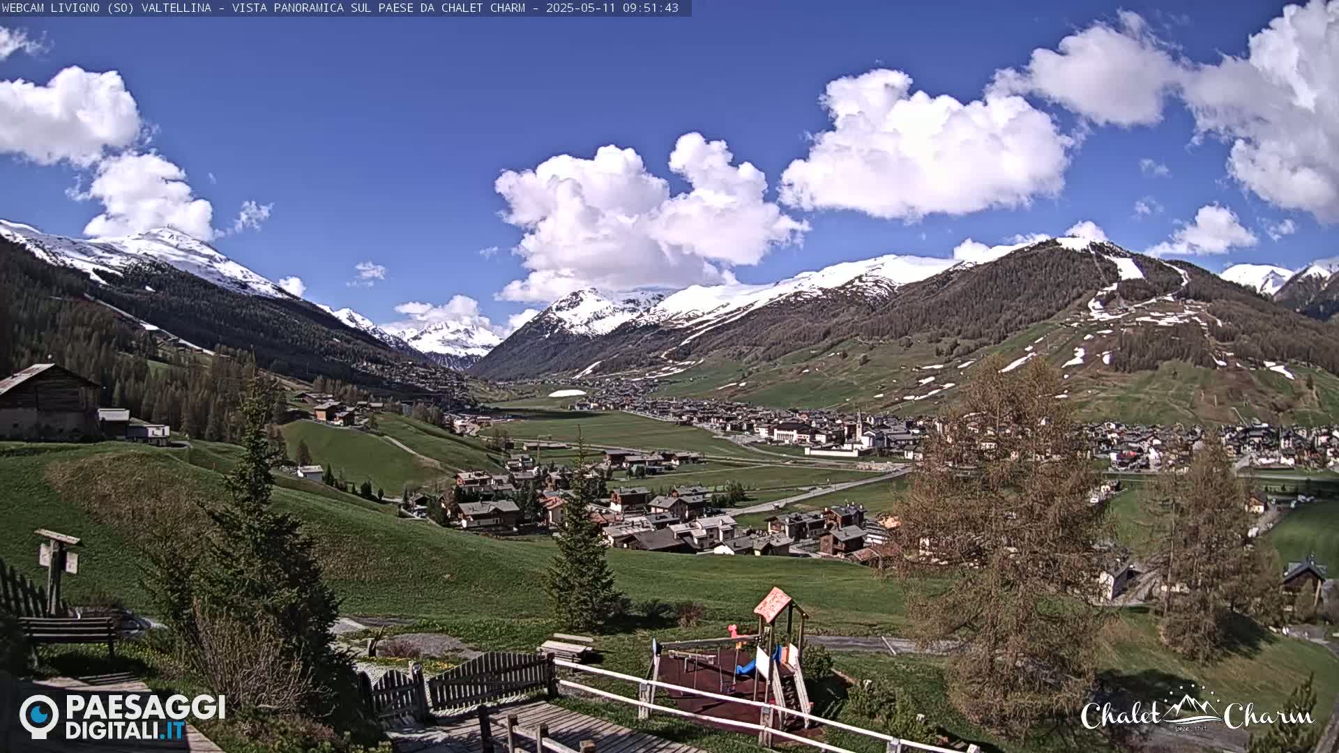 A partly cloudy day reveals a panoramic view of a snow-capped mountain valley with a village nestled below, featuring a playground in the foreground.