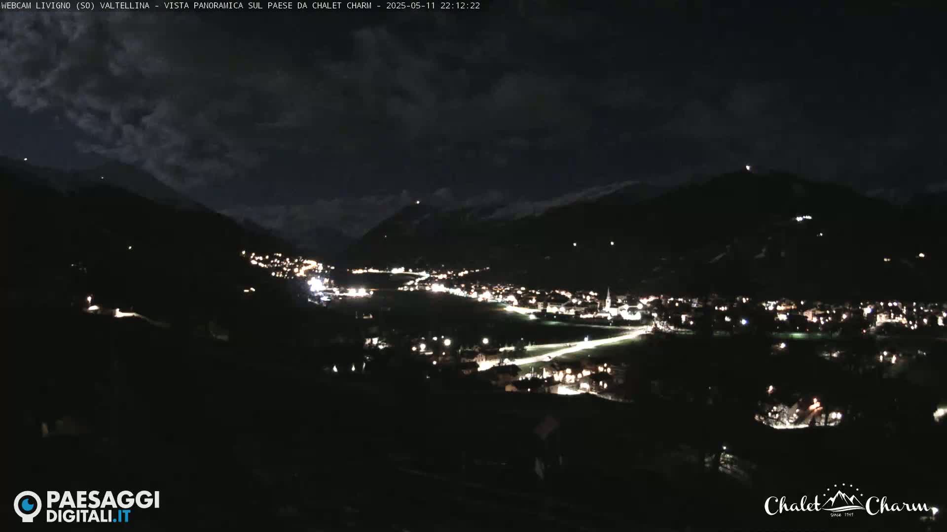 A nighttime, panoramic view of a valley town illuminated by streetlights under a partly cloudy sky.