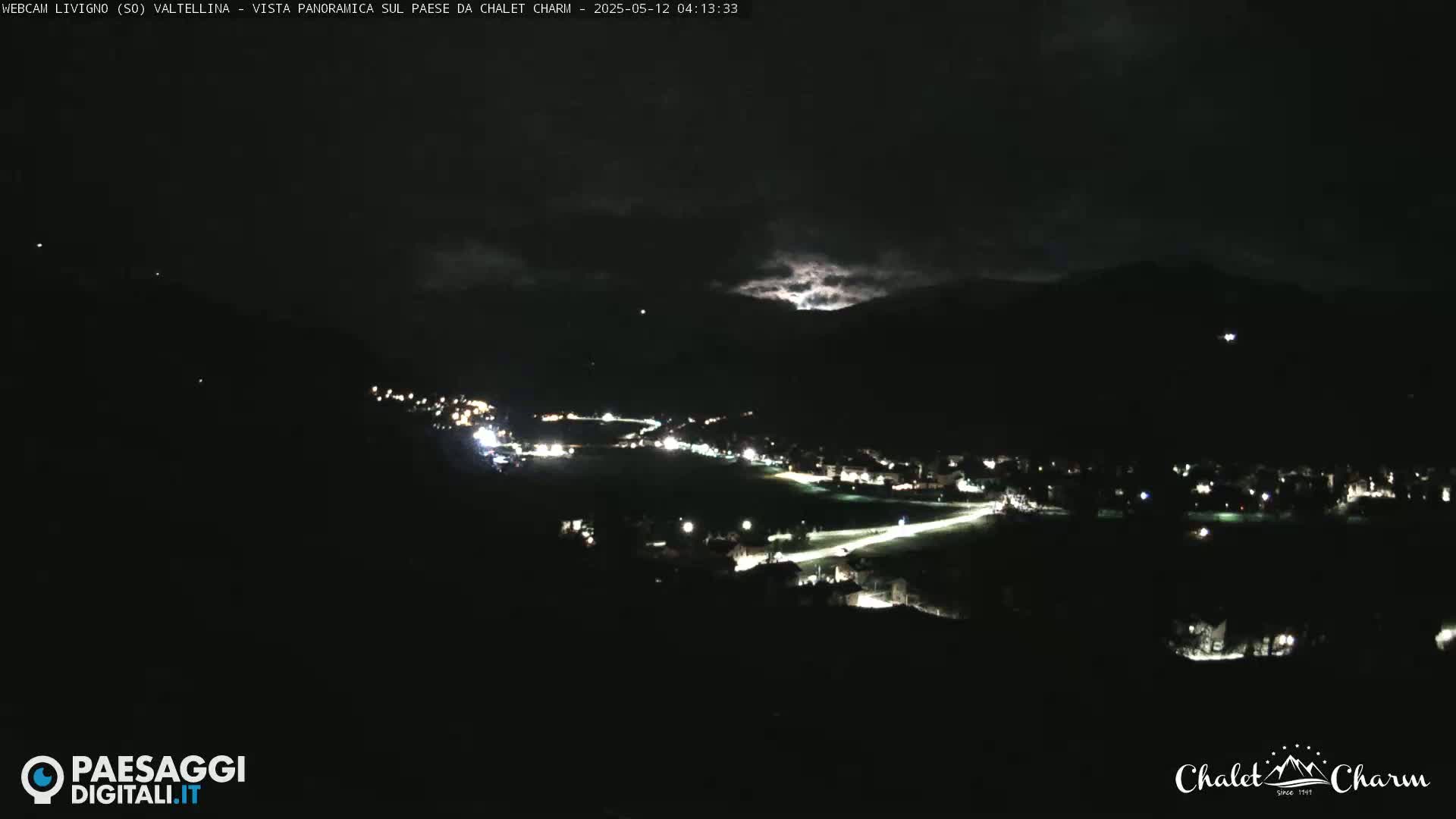 A nighttime, panoramic view of a town nestled in a valley under a partly cloudy sky.