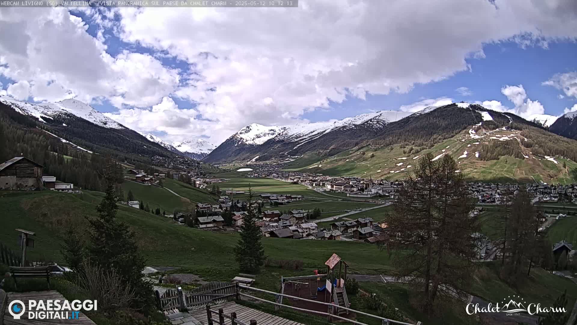 A partly cloudy day reveals a valley town nestled between snow-capped mountains, with a playground visible in the foreground.