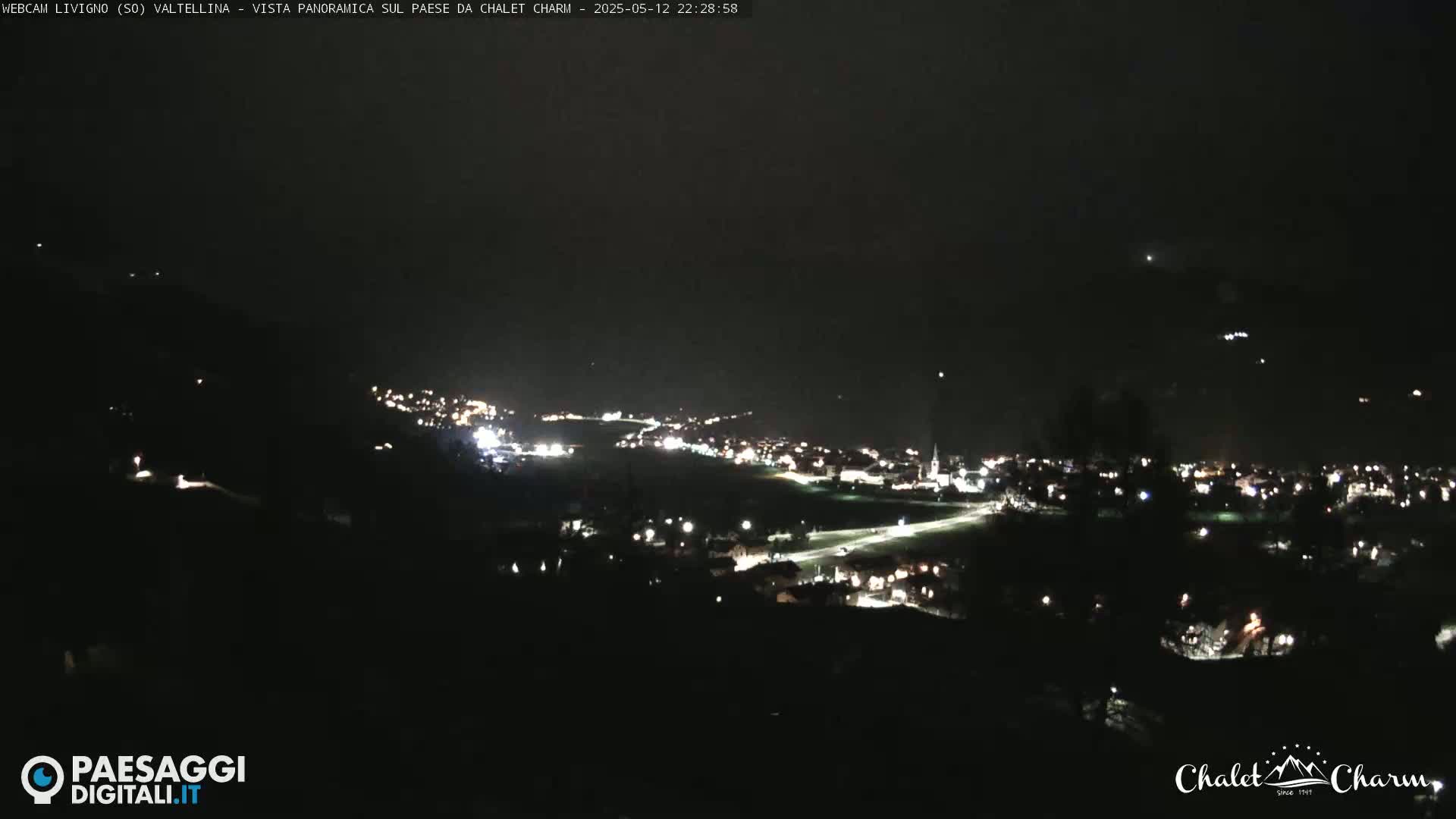 A nighttime, panoramic view shows a valley town illuminated with scattered lights under a dark, clear sky.