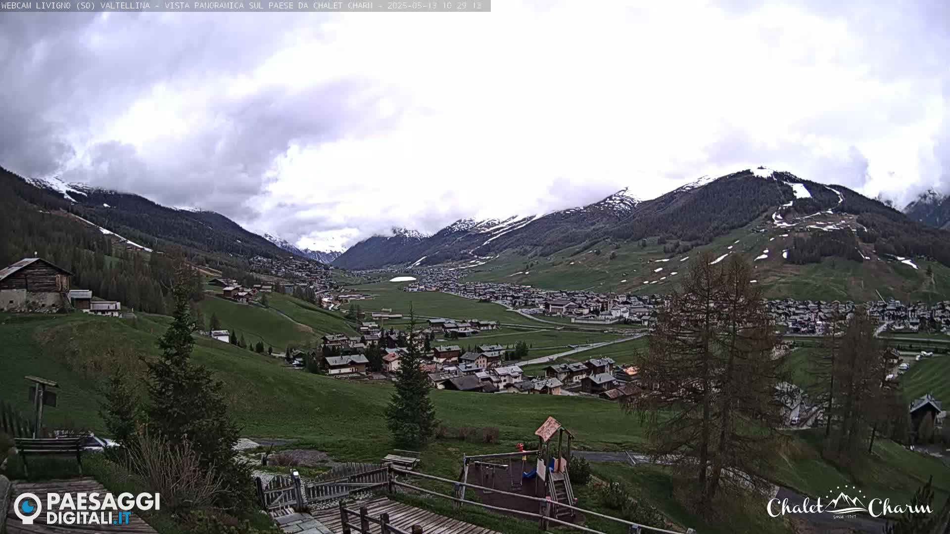 A partially cloudy day reveals a valley town nestled among snow-capped mountains and green hills, with a playground visible in the foreground.