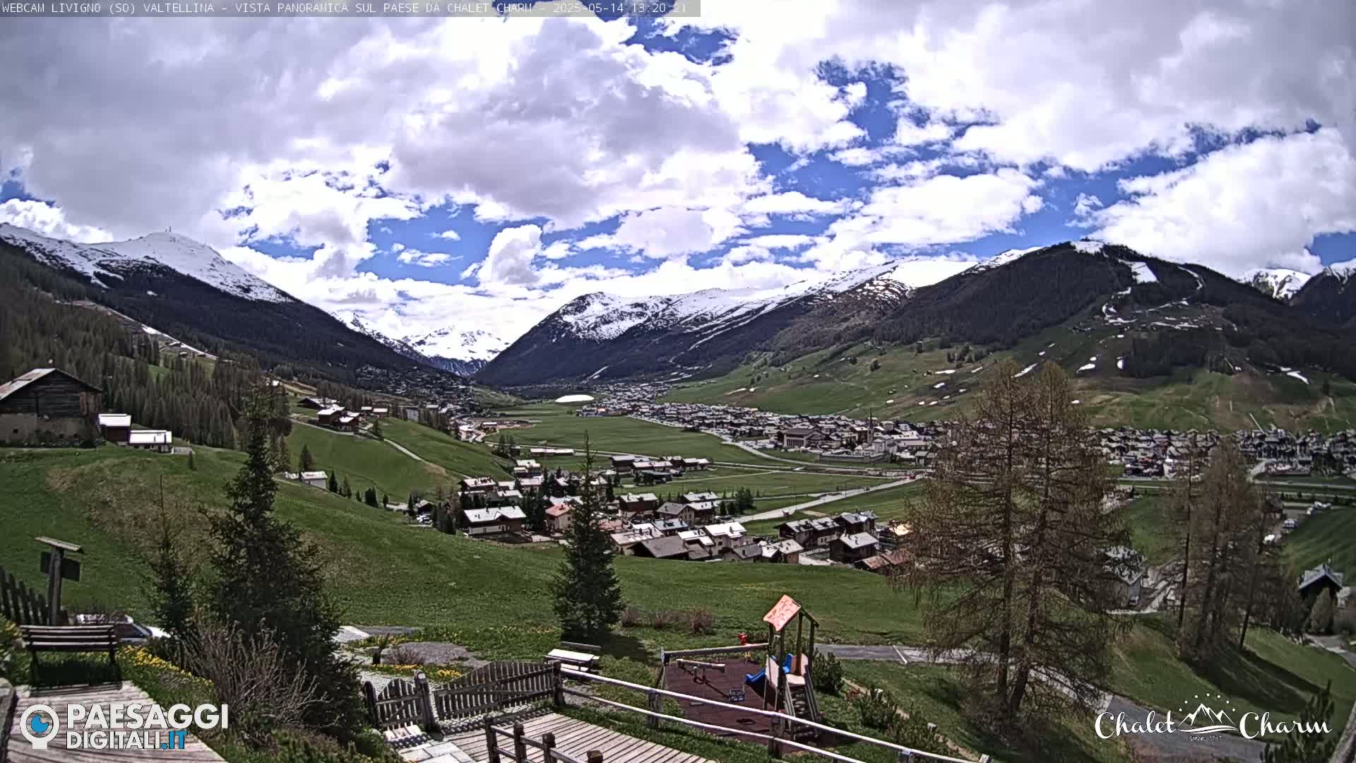 A partly cloudy day reveals a panoramic view of a village nestled in a valley between snow-capped mountains, with a playground visible in the foreground.