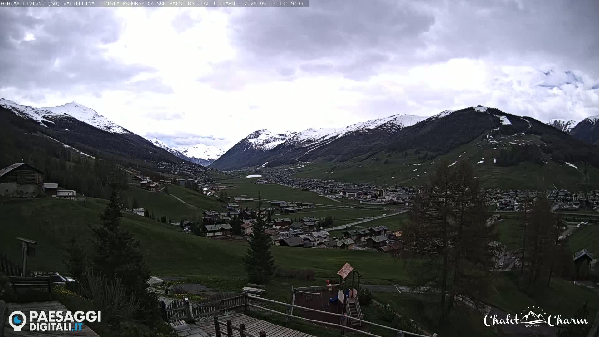 A panoramic view of a snow-capped mountain valley town under a cloudy sky shows a playground in the foreground.