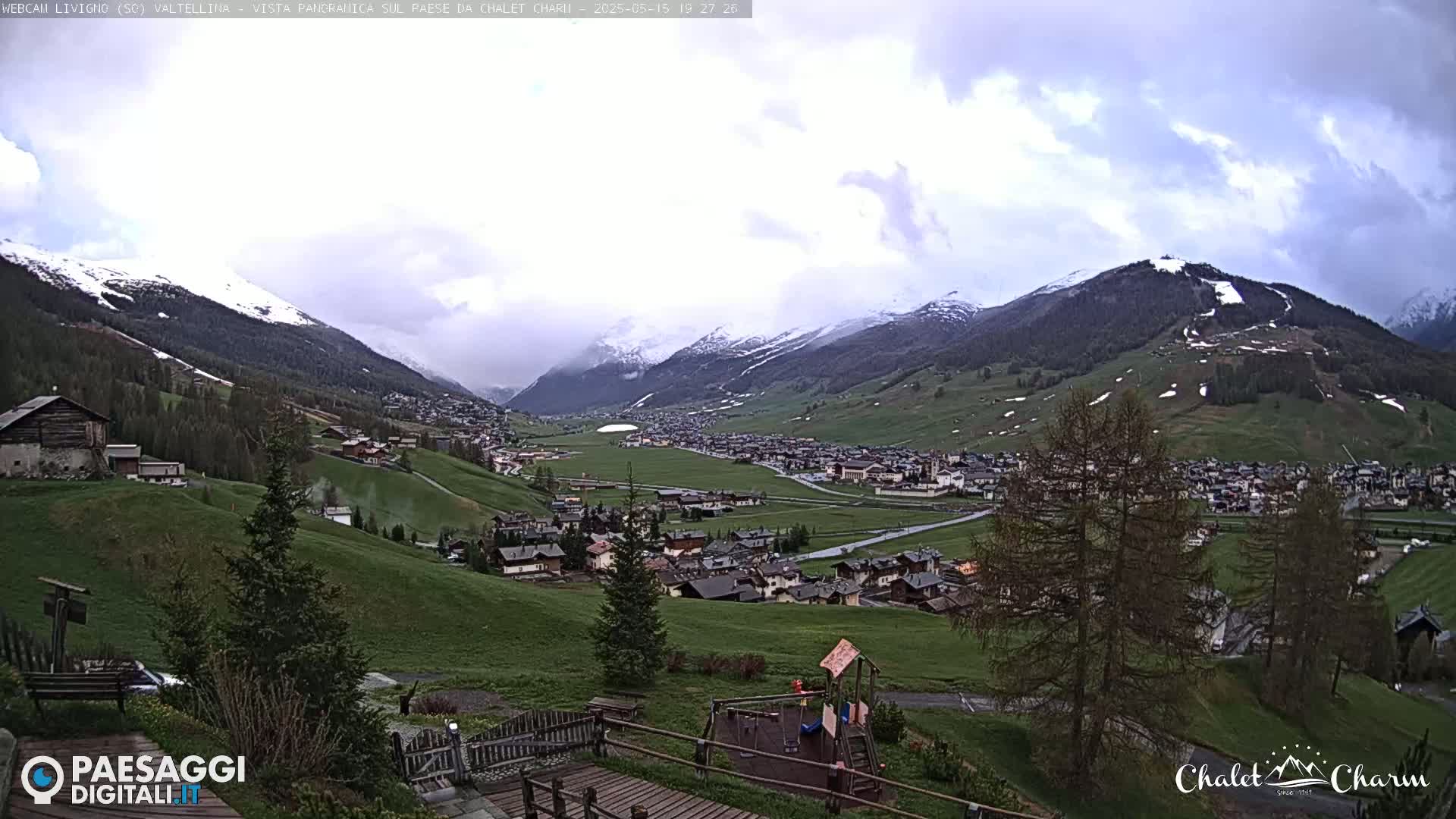 A partly cloudy day reveals a valley town nestled among snow-capped mountains and green hills, with a playground visible in the foreground.