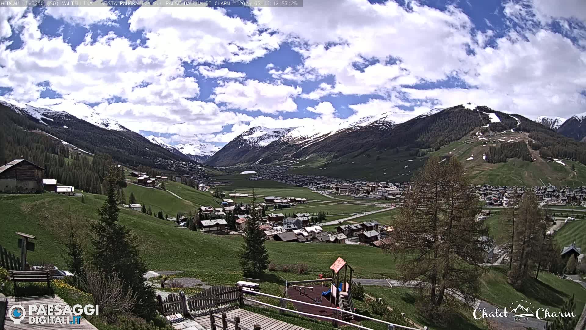 A sunny day with partly cloudy skies overlooks a valley town nestled among snow-capped mountains and green hills, featuring a playground in the foreground.