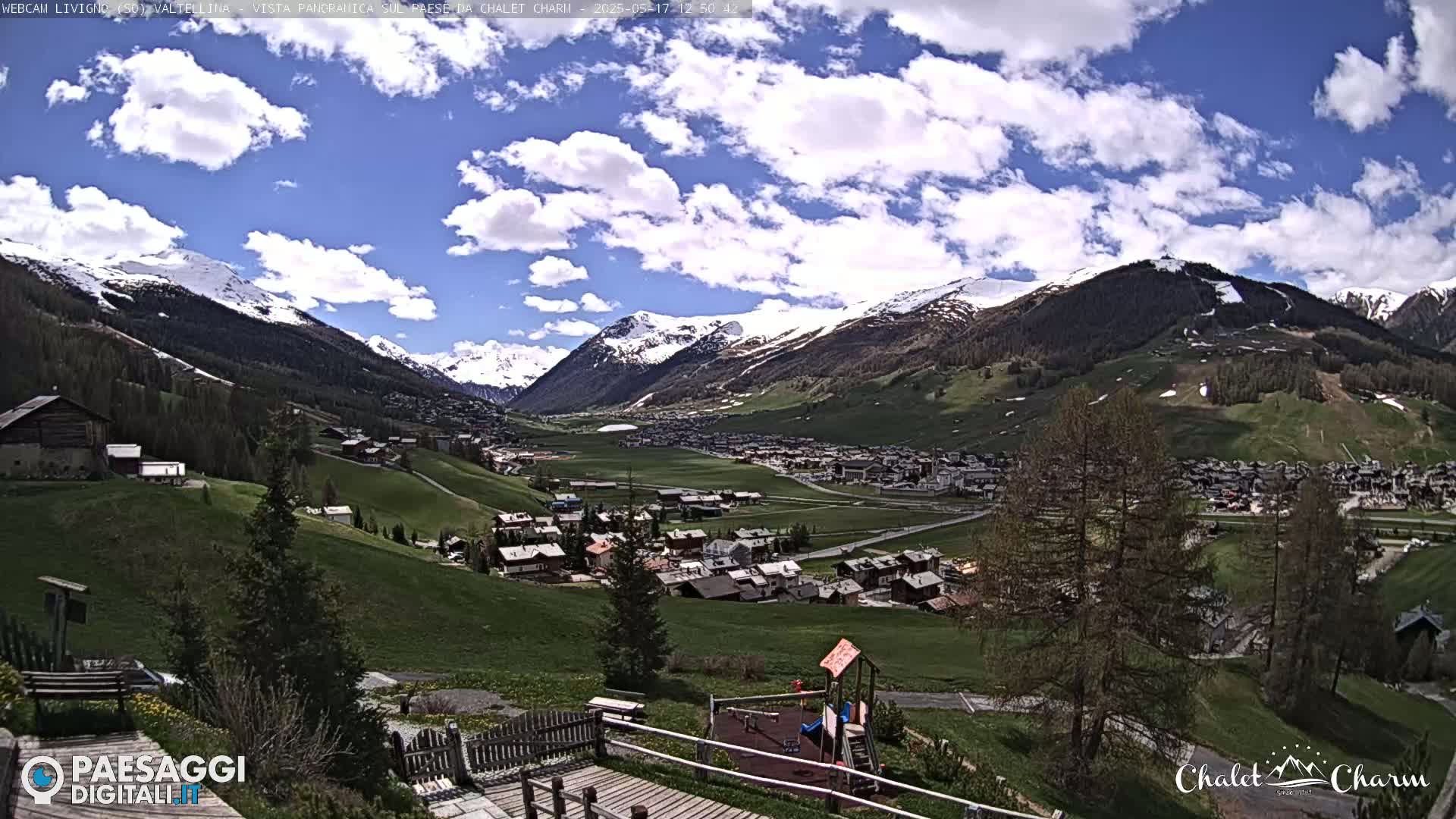 A partly cloudy day reveals a picturesque valley town nestled among snow-capped mountains and green hills, with a playground visible in the foreground.