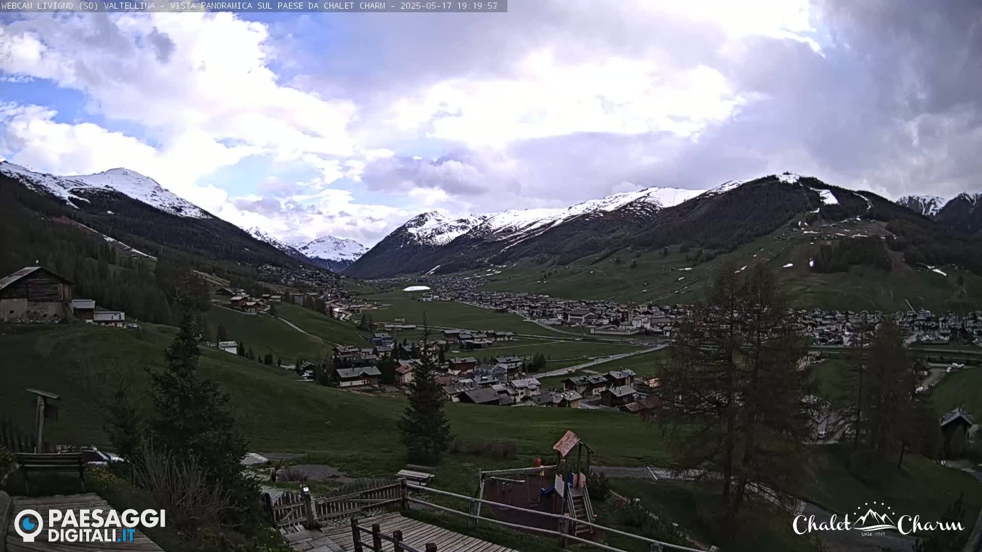 A partly cloudy day reveals a snow-capped mountain range overlooking a valley town with green fields and a playground.