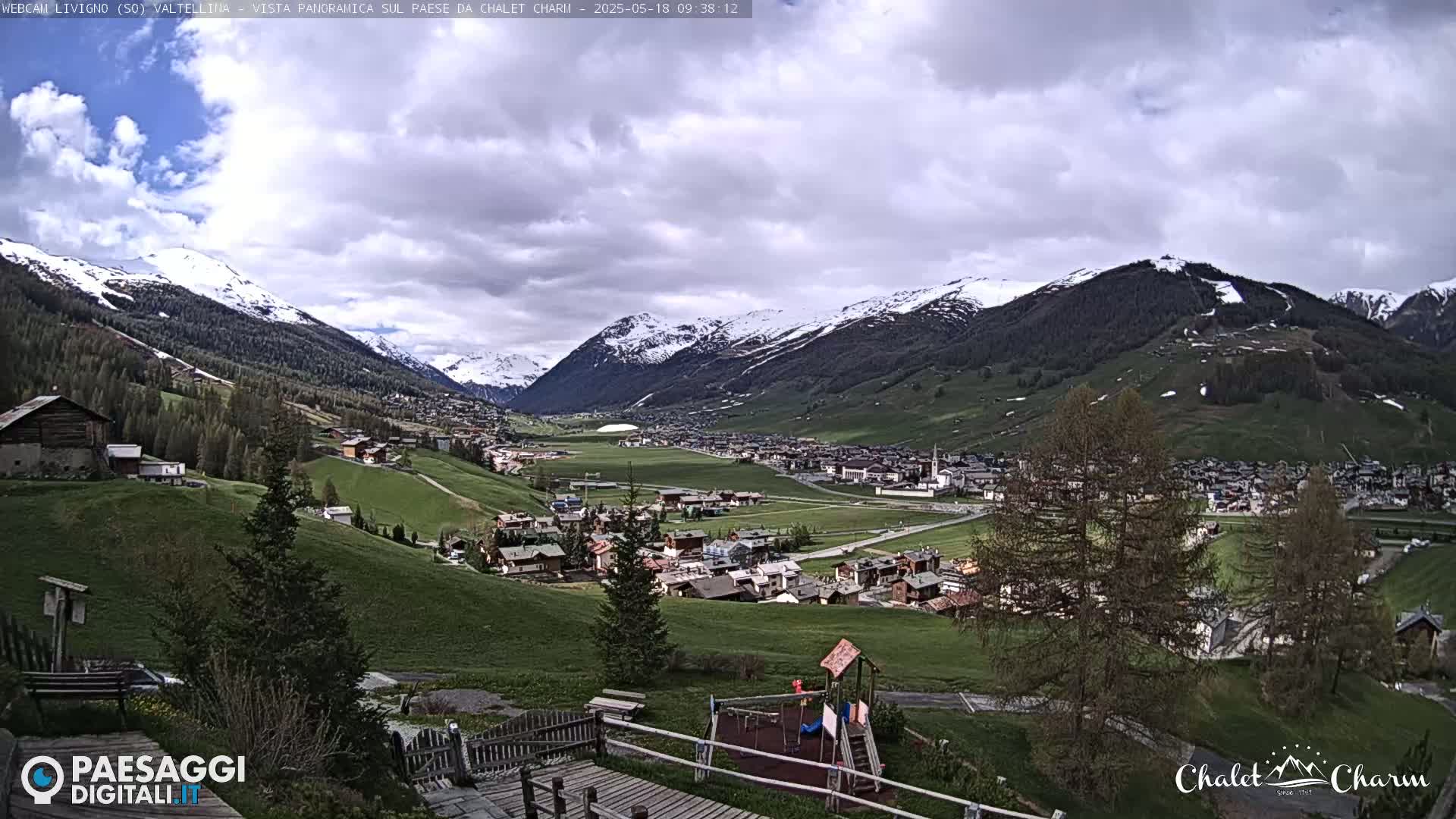 A panoramic view of a village nestled in a valley between snow-capped mountains under a partly cloudy sky.