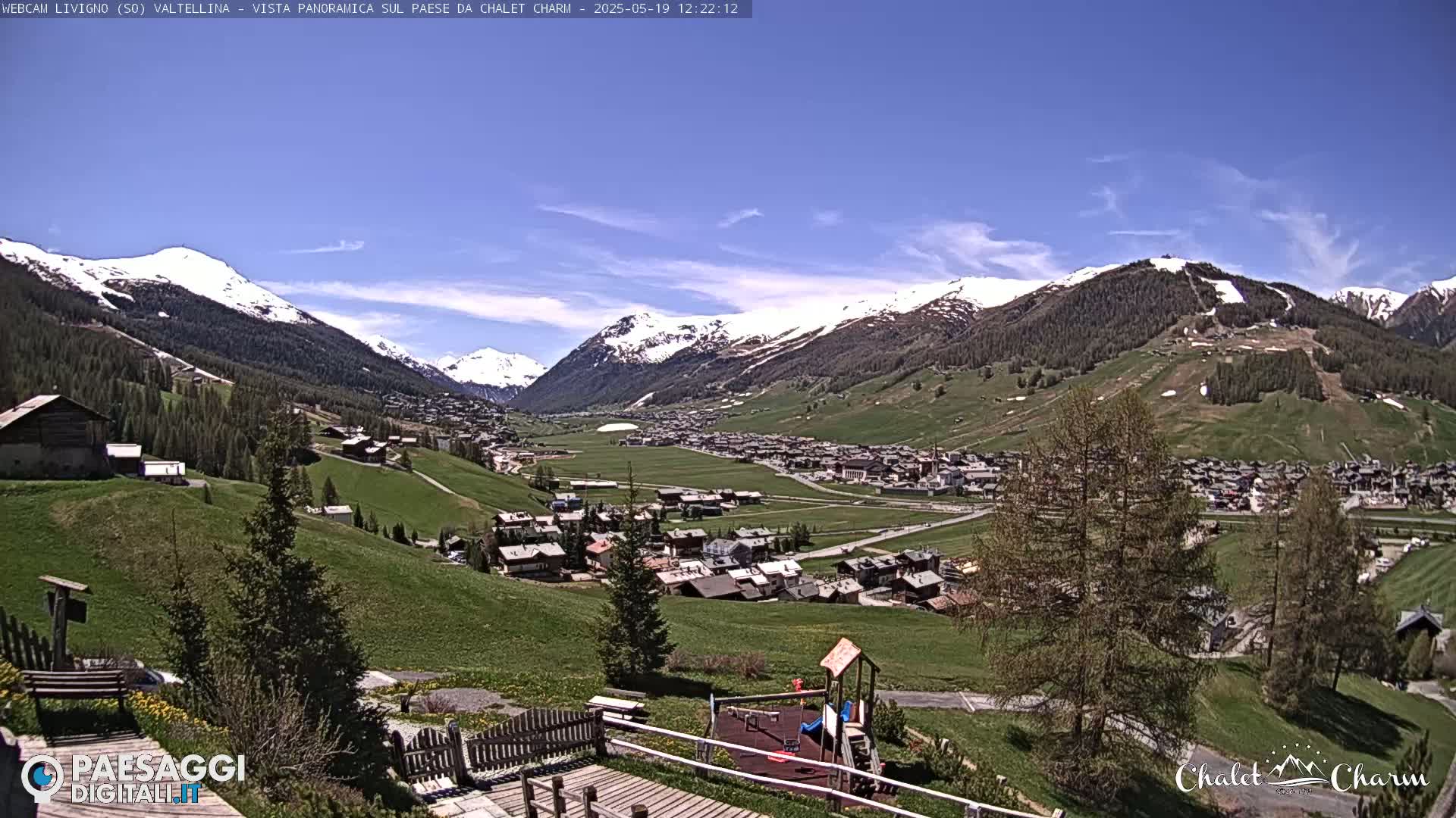 A sunny day reveals a snow-capped mountain range overlooking a valley town with green hills and a playground.