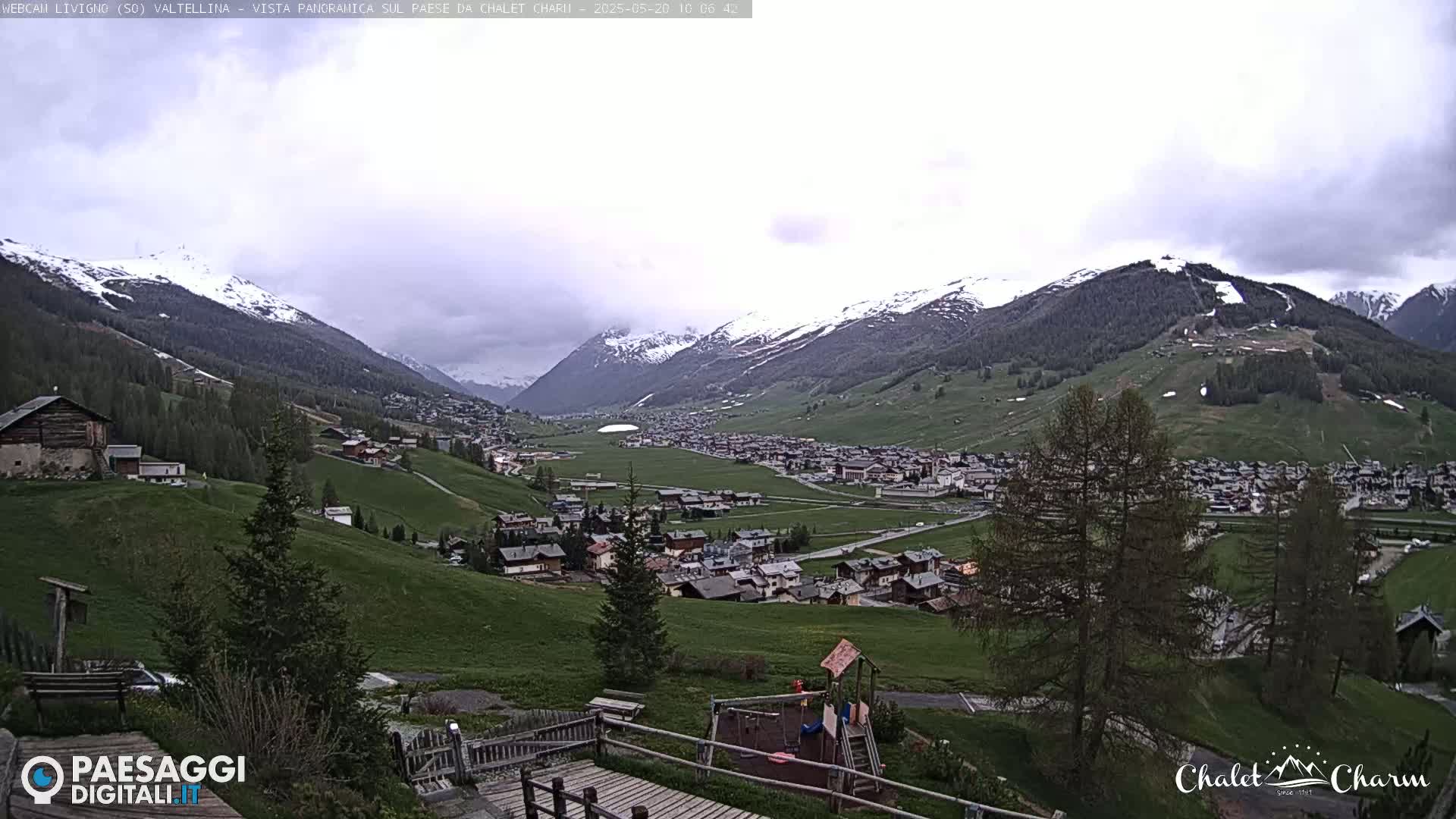 A partly cloudy day reveals a valley town nestled among snow-capped mountains and green hills, with a playground visible in the foreground.