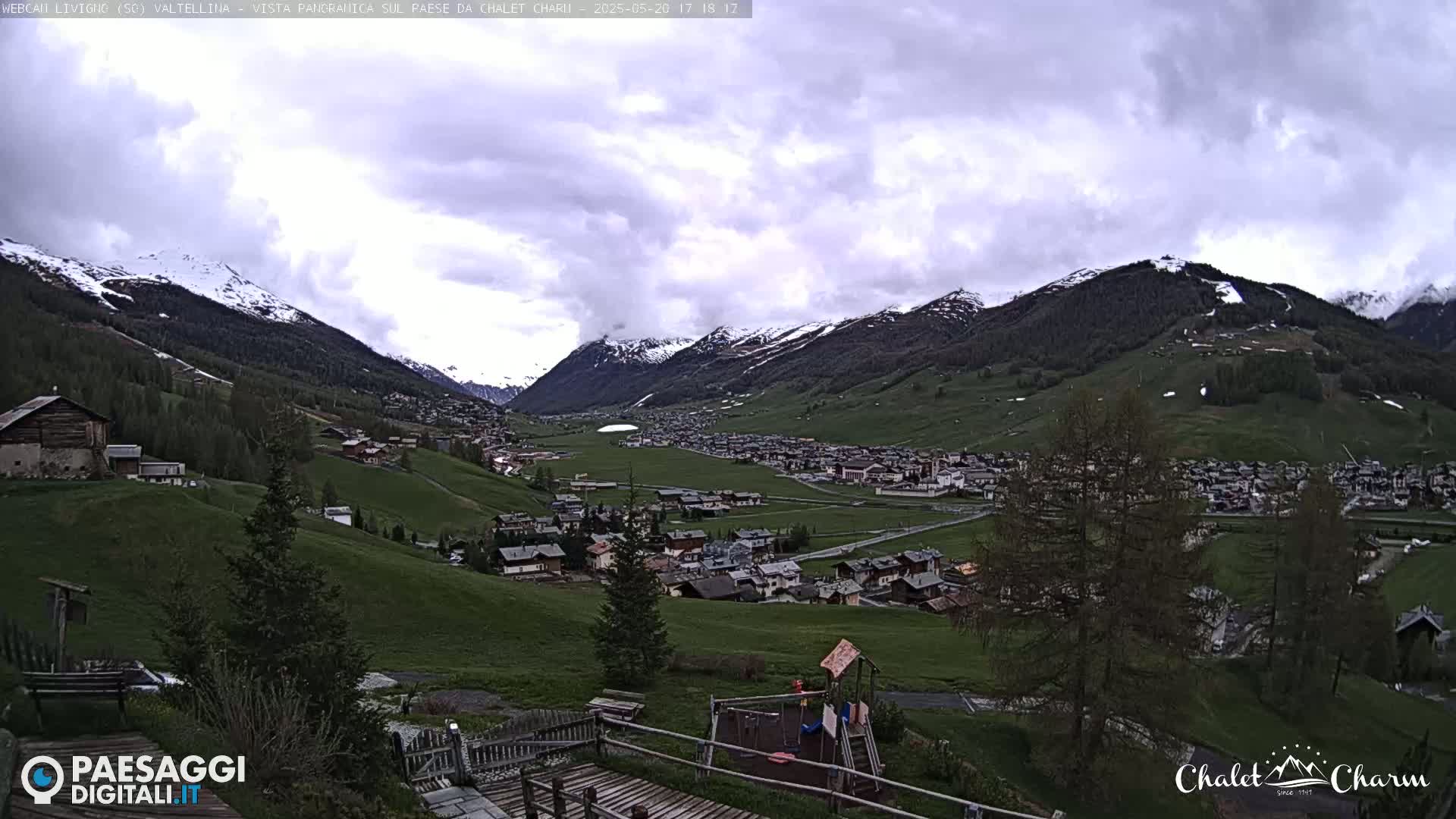 A panoramic view of a village nestled in a valley between snow-capped mountains under a cloudy sky.
