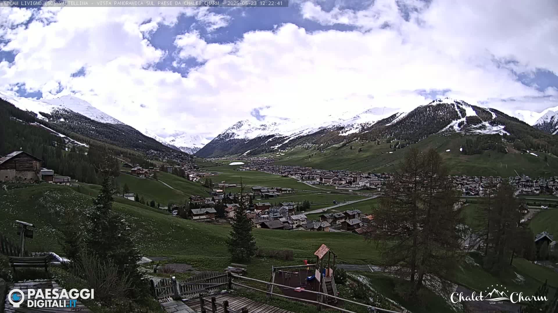 A partly cloudy day reveals a snow-capped mountain range overlooking a valley town with green pastures and a playground.