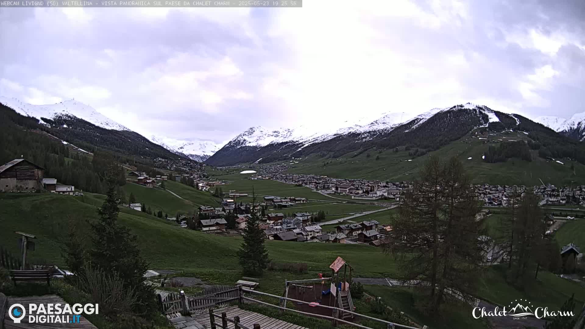 A picturesque mountain village nestled in a valley, with snow-capped mountains in the background under a partly cloudy sky.