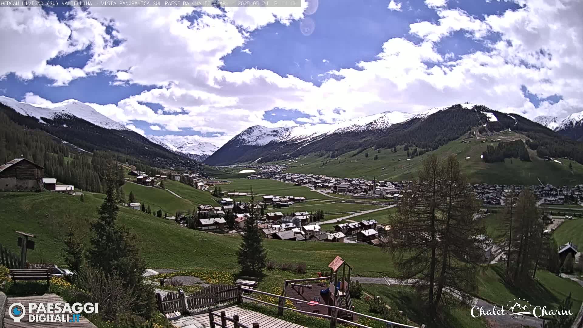 A partly cloudy day reveals a picturesque valley town nestled among snow-capped mountains and green hills.