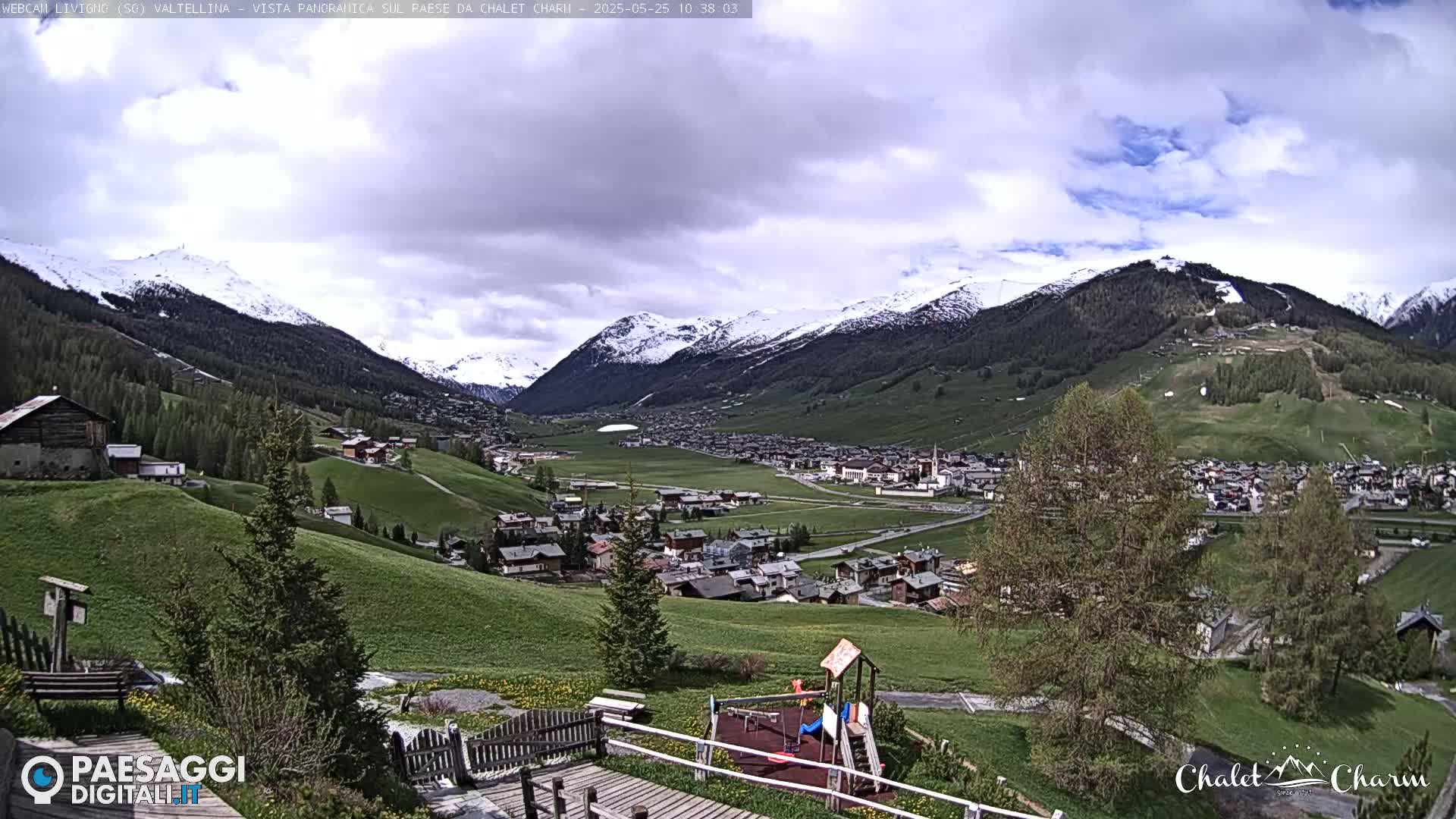 A partly cloudy day reveals a mountain valley town nestled amongst snow-capped mountains and green hills, with a small playground visible in the foreground.